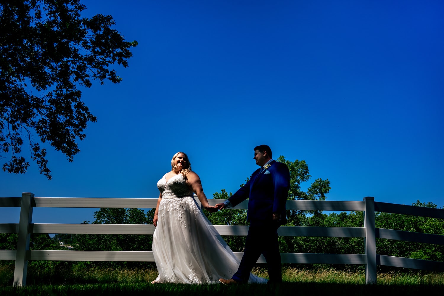 A colorful, candid picture of a bride leading her groom across a bright green yard on their summer wedding day at The Farmhouse KC Event Venue. 