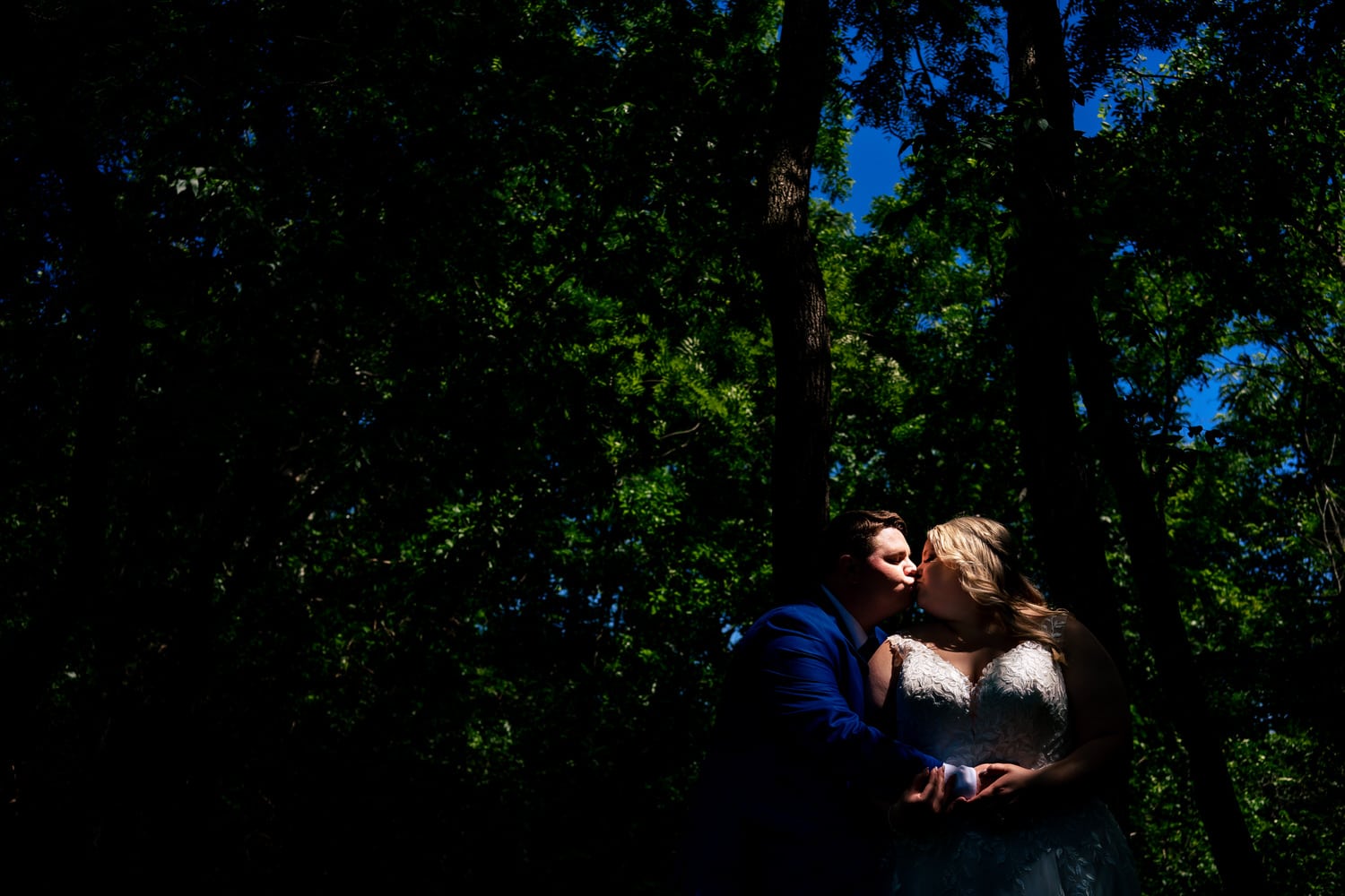 A colorful, candid picture of a bride and groom sharing an embrace, a colorful canopy of blue sky and green leaves visible behind them. 