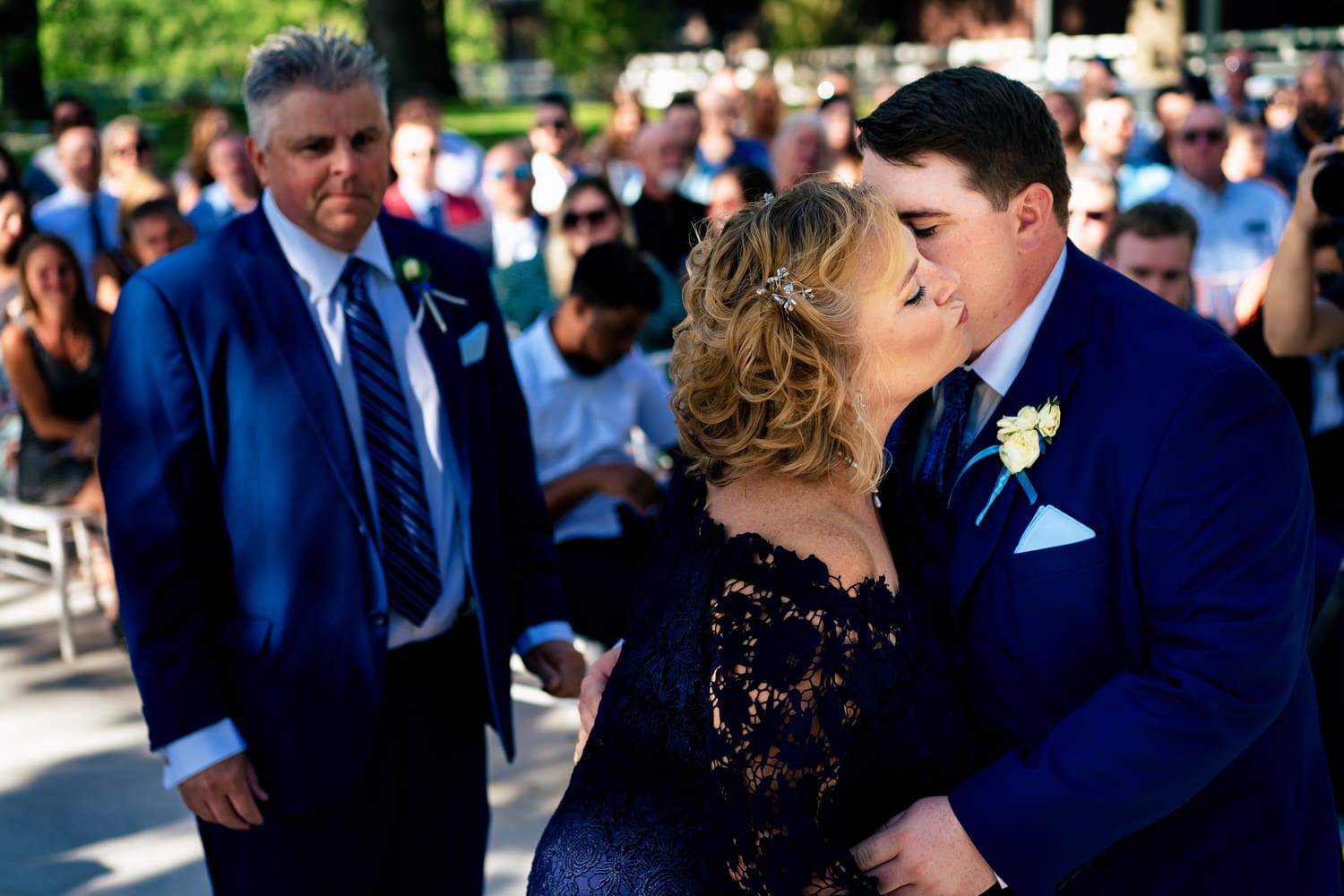 A colorful, candid picture of a groom kissing his mom as she sits down on a summer wedding day at The Farmhouse KC Event Venue. 