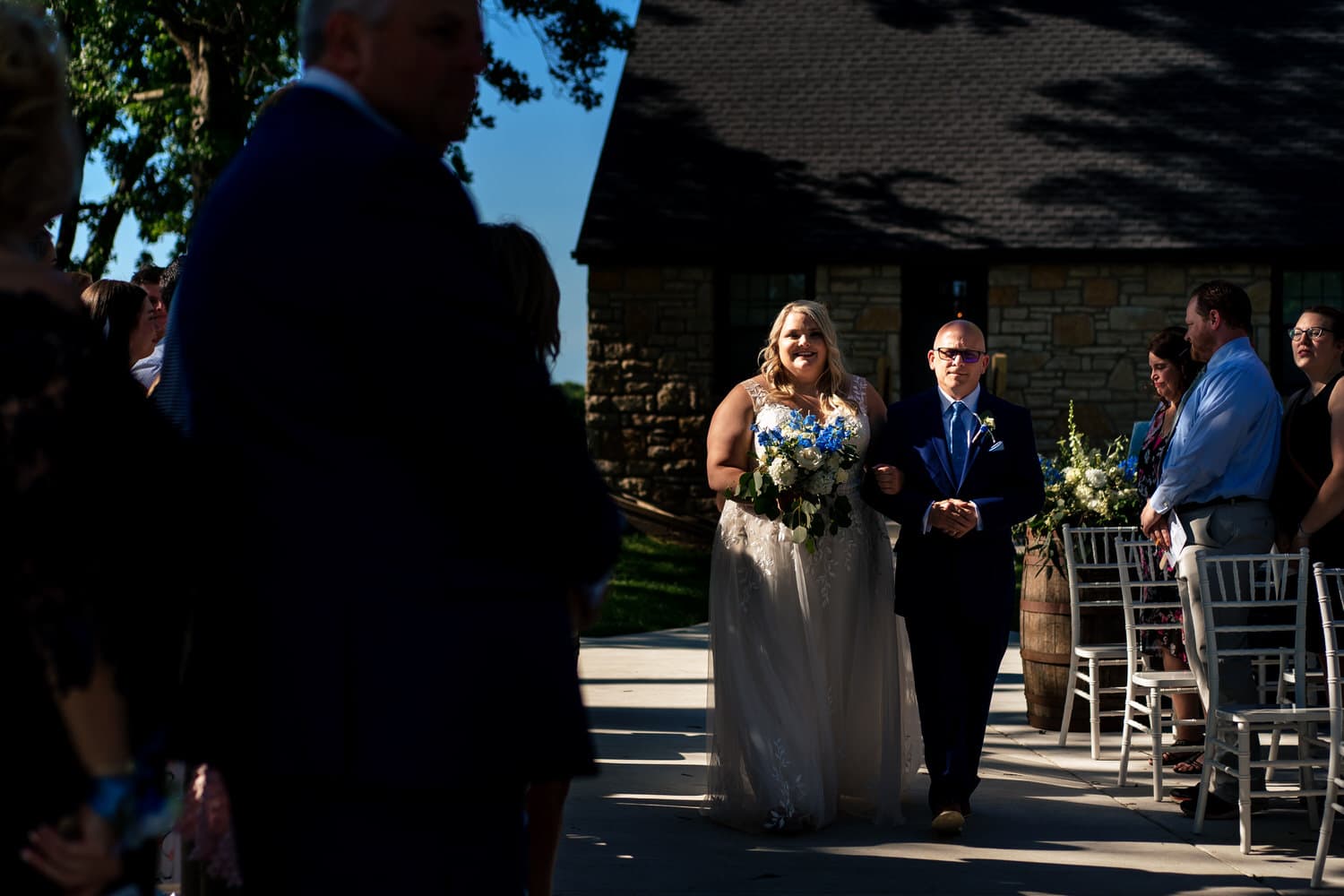 A colorful picture of a bride and her father walking down the aisle on a summer wedding day at The Farmhouse KC Event Venue. 