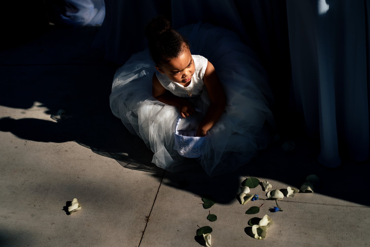 A candid picture taken from the top looking down of a flower girl in a white dress squatting on the cement during a wedding ceremony at The Farmhouse KC Event Venue. 
