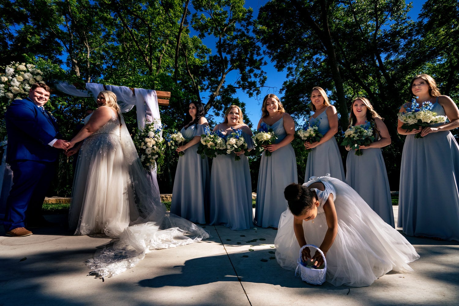 A candid picture taken from the top looking down of a flower girl in a white dress squatting on the cement during a wedding ceremony at The Farmhouse KC Event Venue. 