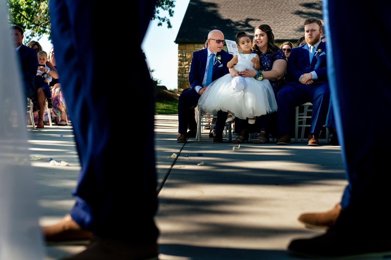 A colorful picture taken between the legs of groomsmen of a bride's dad, mom, and flower girl during a summer wedding ceremony at The Farmhouse KC Event Venue in Kansas City. 