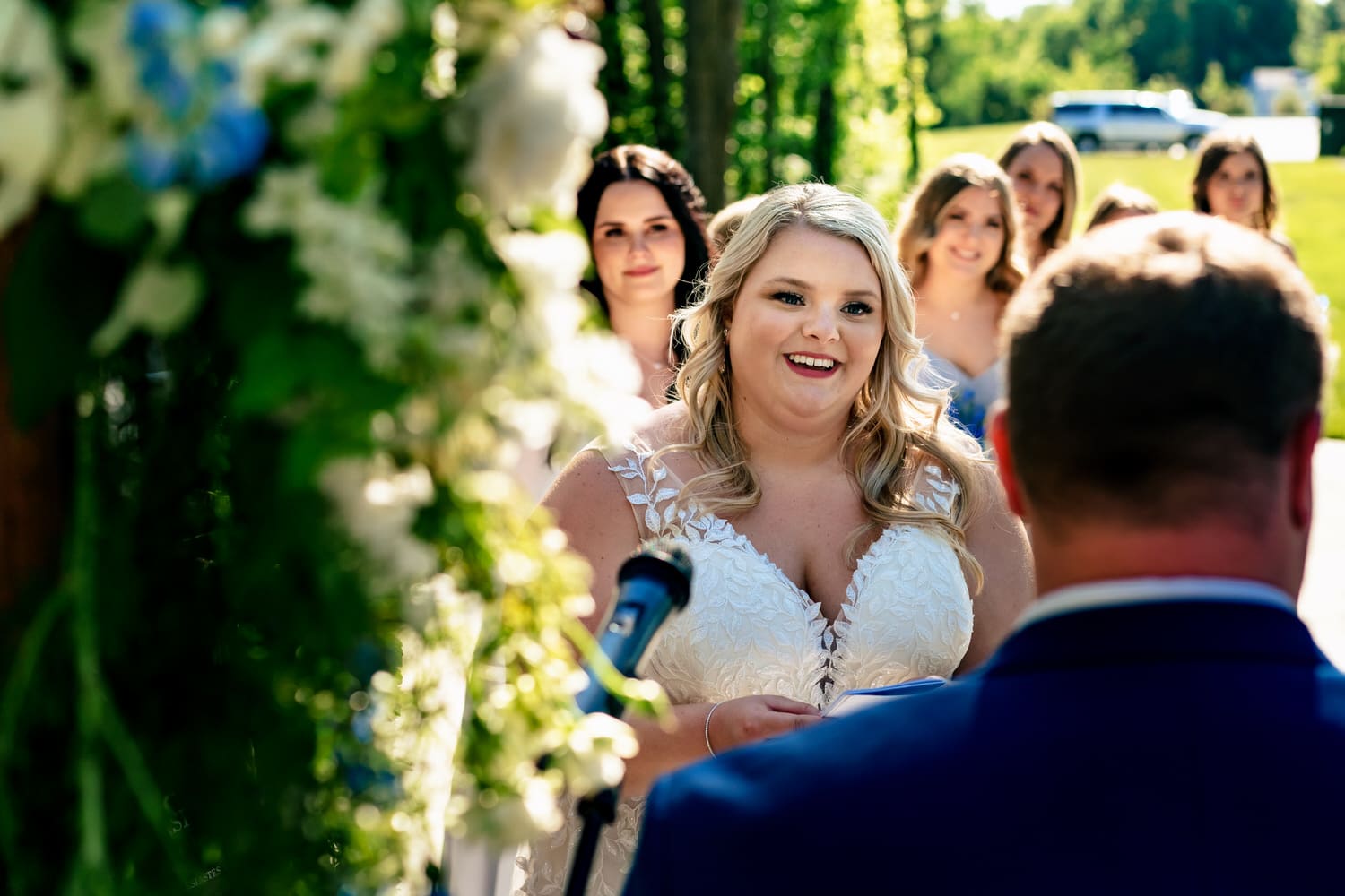 A candid picture of a bride smiling as she reads her wedding vows to her groom during a summer wedding ceremony at The Farmhouse KC Event Venue. 