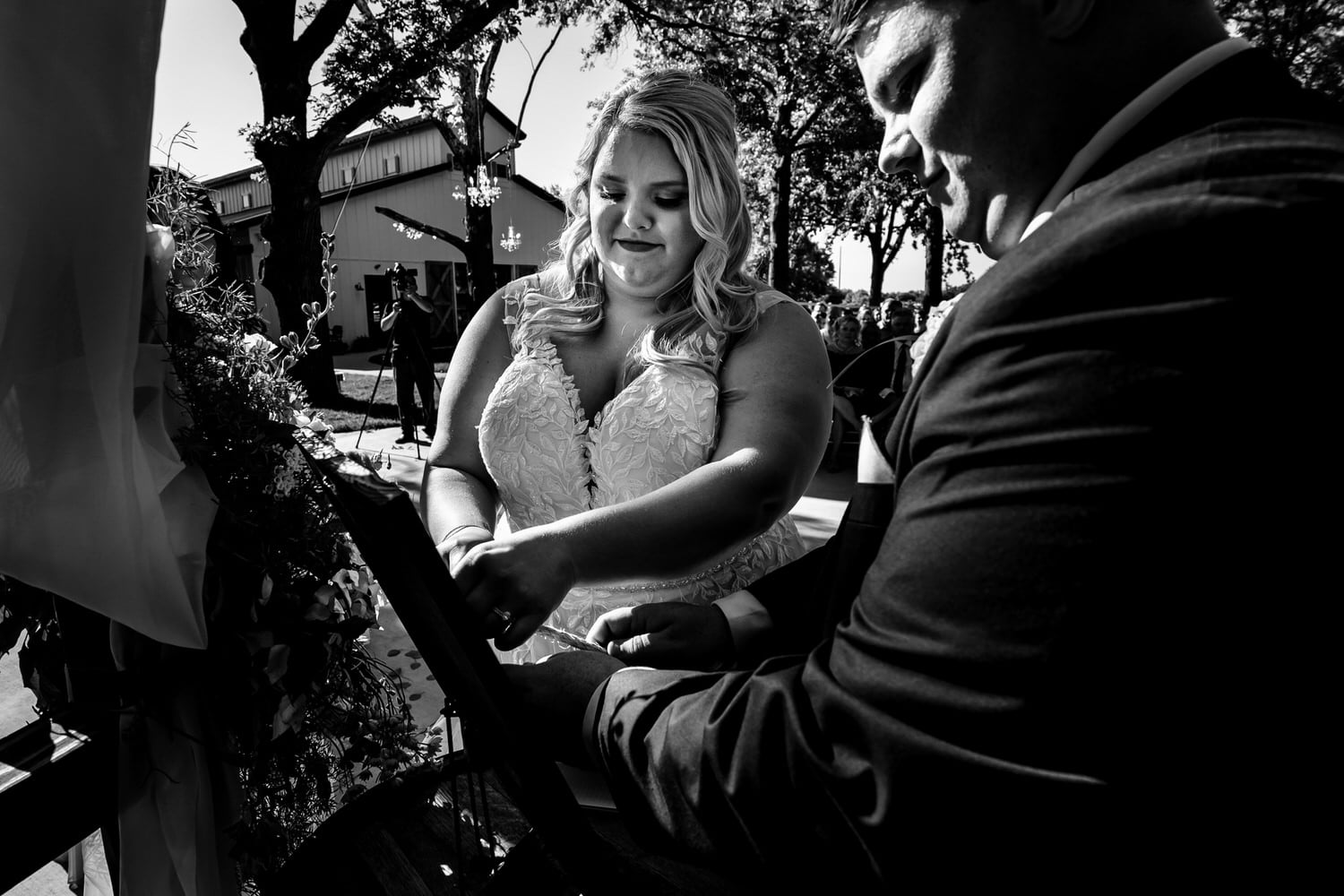 A candid, black and white picture of a bride and groom tying a rope of three cords during an outside summer wedding ceremony at The Farmhouse KC Event Venue. 