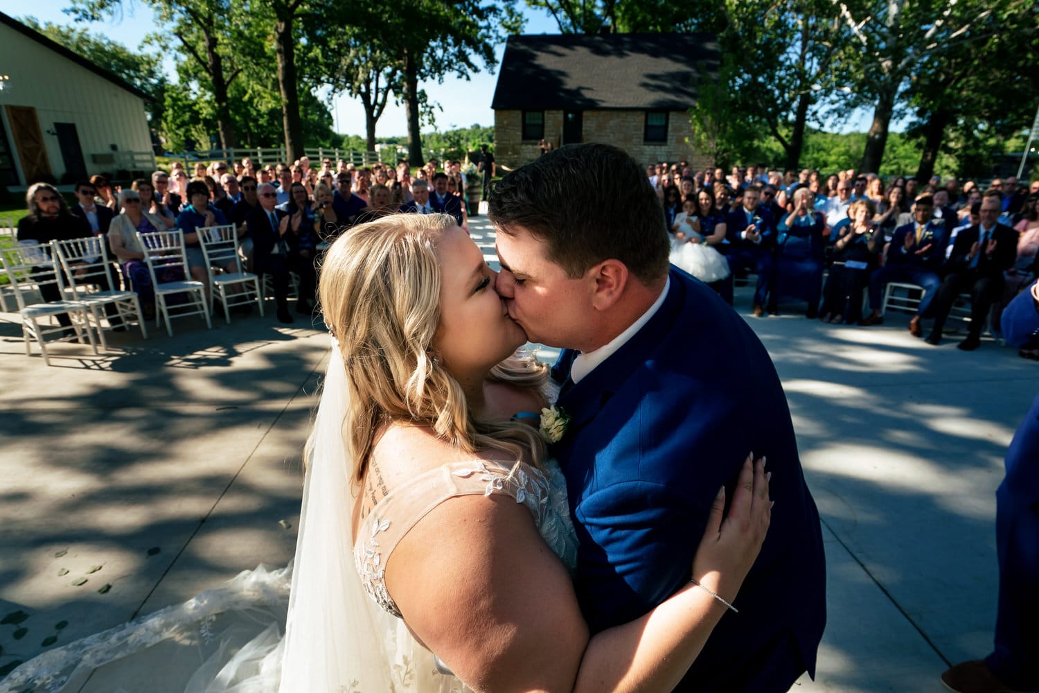 A candid picture taken from behind the altar of a bride and groom sharing a first kiss as their family and friends clap in celebration behind them during a summer wedding ceremony at The Farmhouse KC Event Venue. 