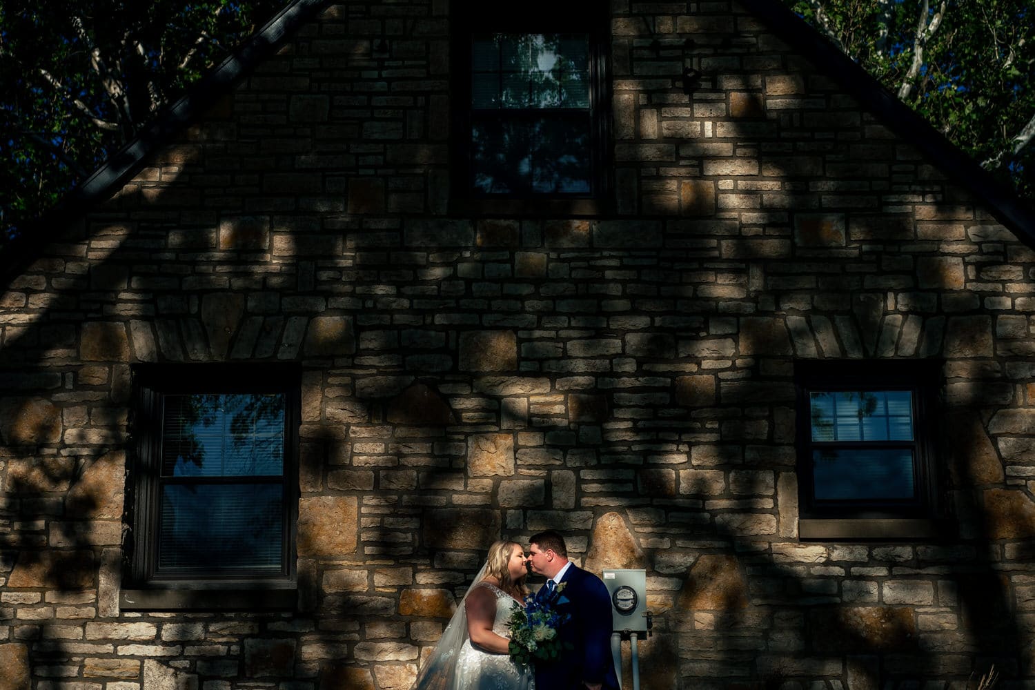 A wide, colorful picture of a bride and groom holding hands, standing in front of the side of a stone building looking away from each other on their summer wedding day at The Farmhouse KC Event Venue. 