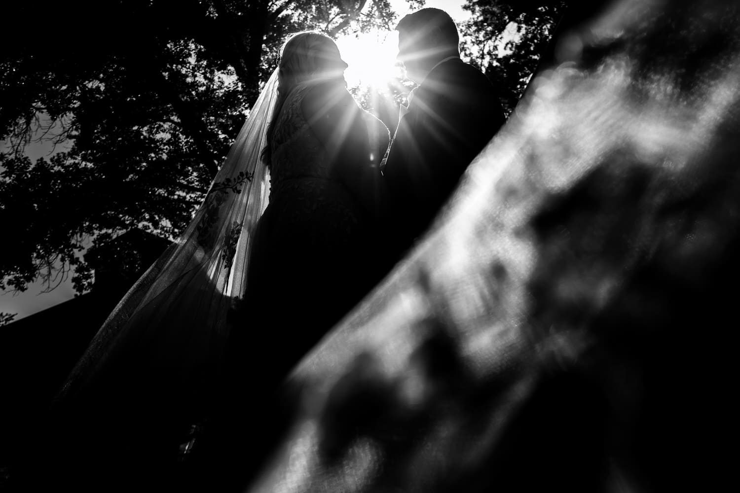 A dramatic black and white portrait taken from the ground looking up of a bride and groom sharing an embrace, the bride's veil wrapped around them on a summer wedding day at The Farmhouse KC Event Venue. 