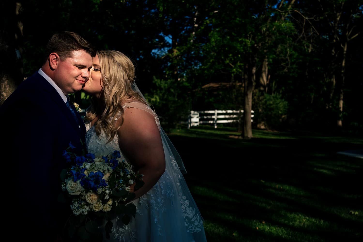 A colorful, candid picture of a bride leaning in to kiss her groom on the cheek on a summer wedding day at The Farmhouse KC Event Venue. 