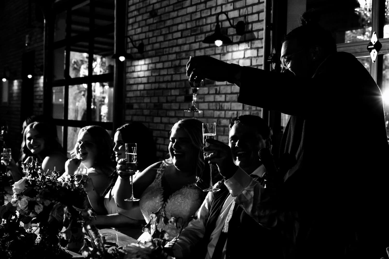 A dramatic black and white picture of a groomsman raising his champagne flute in a toast to the bride and groom during a summer wedding reception at The Farmhouse KC Event Venue. 