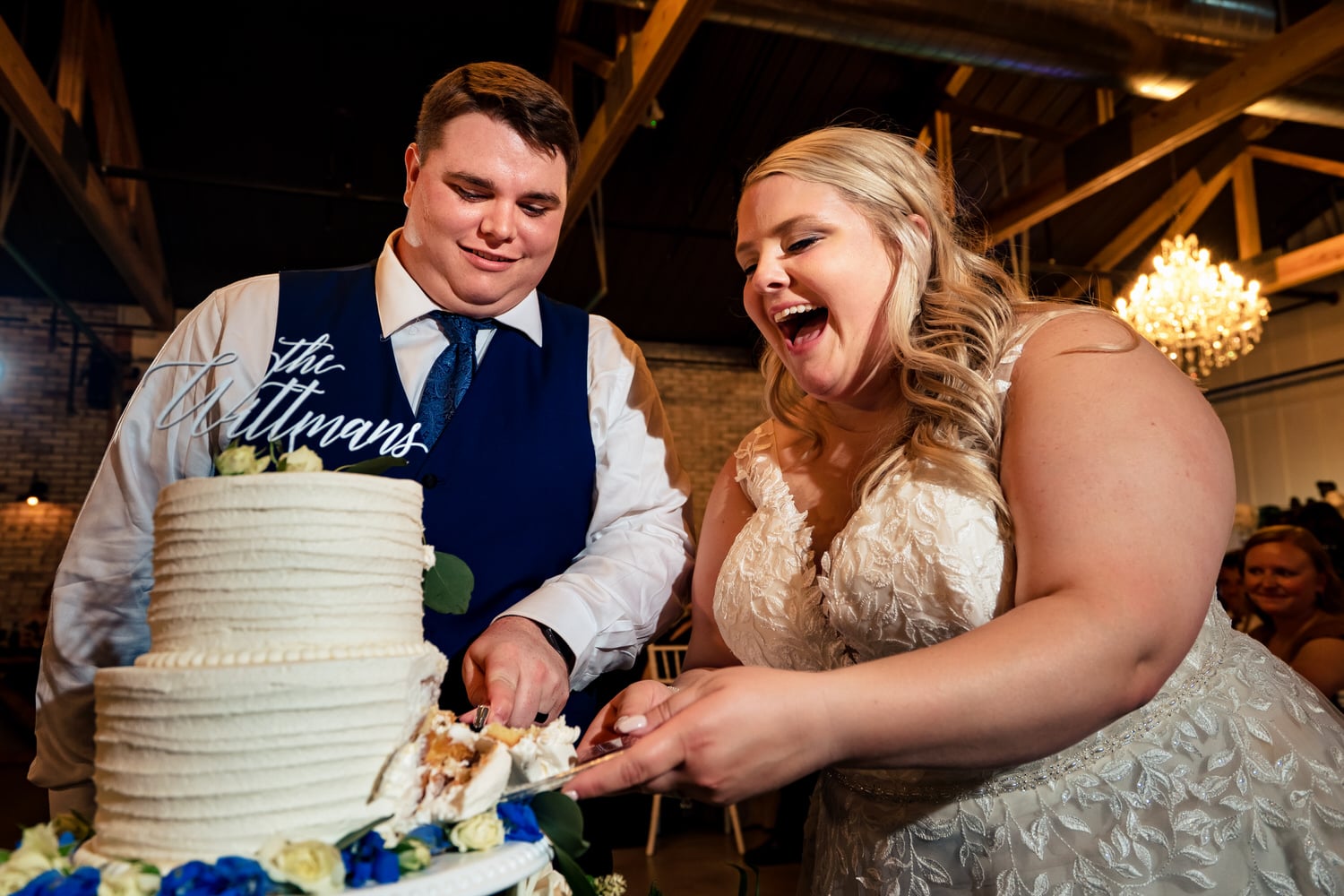 A candid, colorful picture of a bride and groom laughing as they cut a white wedding cake during their summer wedding reception at The Farmhouse KC Event Venue. 