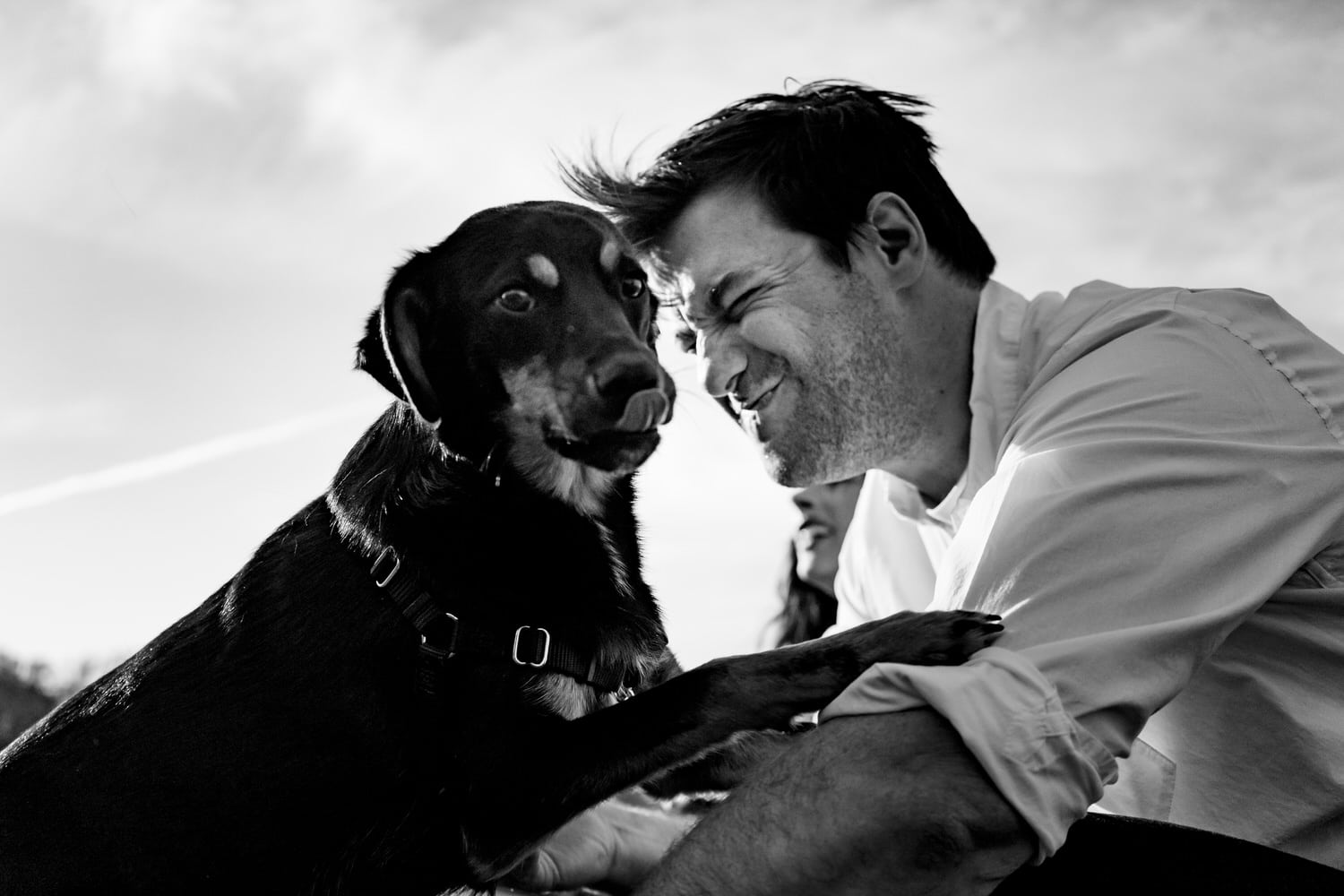 A candid black and white picture of a dog licking his tongue seconds after licking a man's face as the man grimaces in the background. 