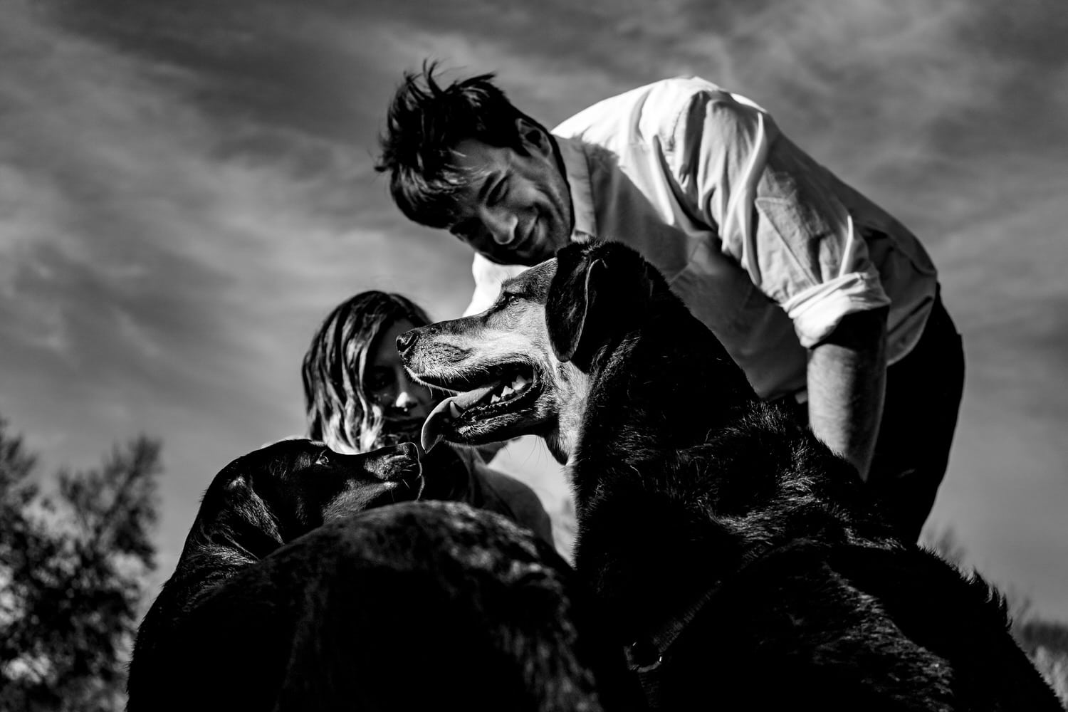 A candid black and white picture of a man and woman bent down petting two dogs during their spring engagement session at Loose Park in Kansas City. 