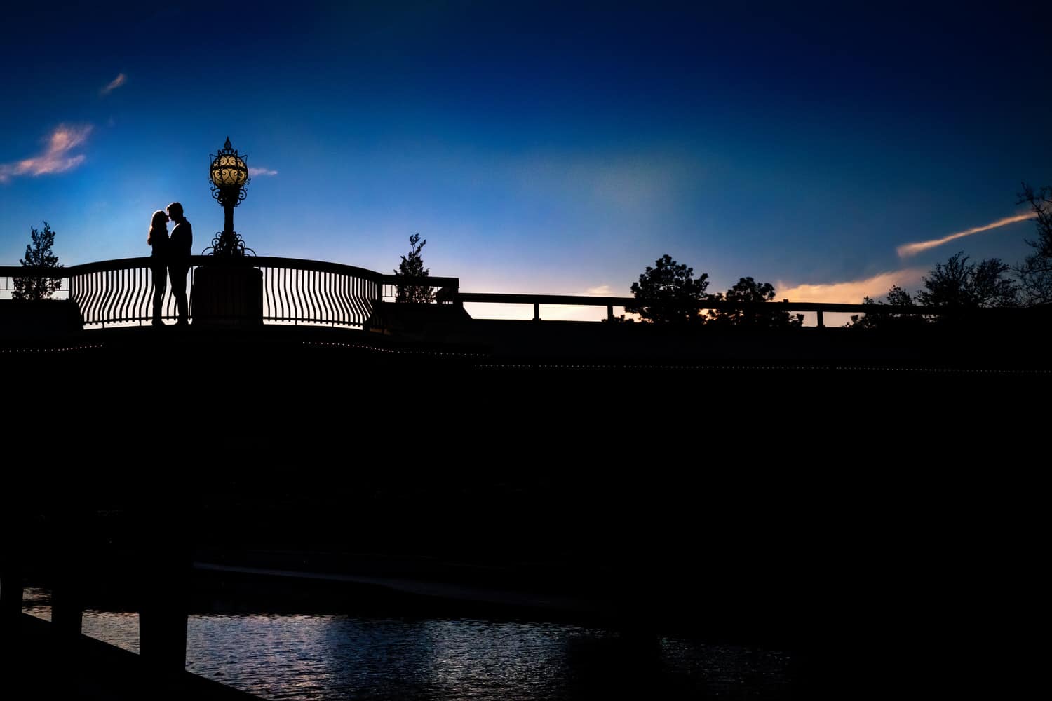 A colorful, silhouette of a couple standing on a bridge over Brush Creek in Kansas City during the engagement session. 