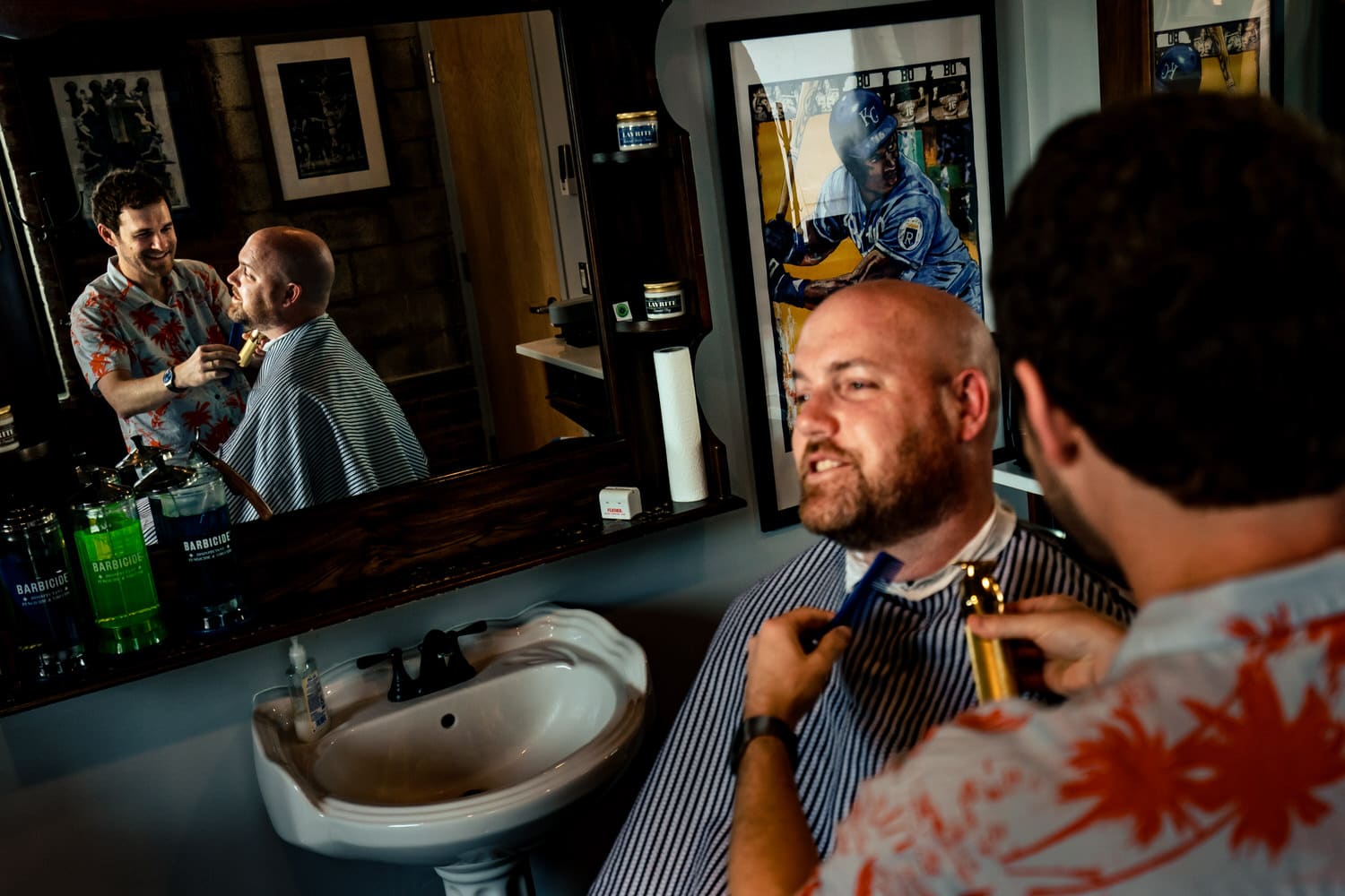 A candid, colorful picture of a man getting his beard trimmed, his reflection visible in the mirror behind him on the morning of his summer wedding day in Kansas City. 