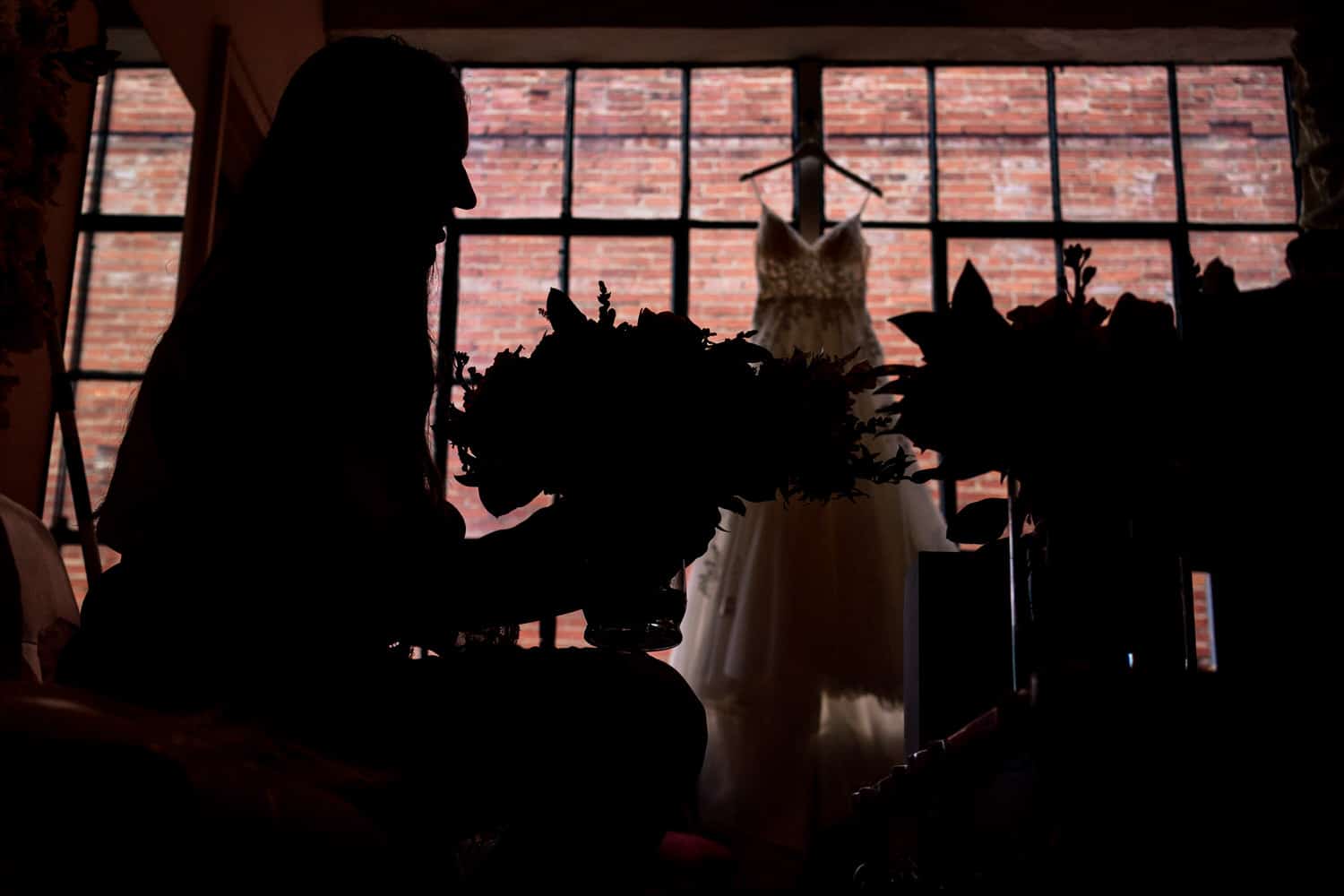 A colorful silhouette of a woman holding a bouquet of flowers, a wedding gown hanging in a window visible behind her on her summer wedding day at The Bride and The Bauer in Kansas City. 