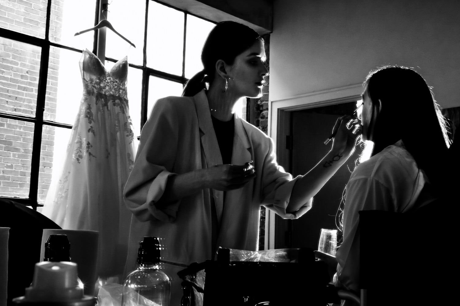 A candid black and white picture of a makeup artist applying makeup to a bride, her wedding gown visible in the window behind her on a summer wedding day at The Bauer. 