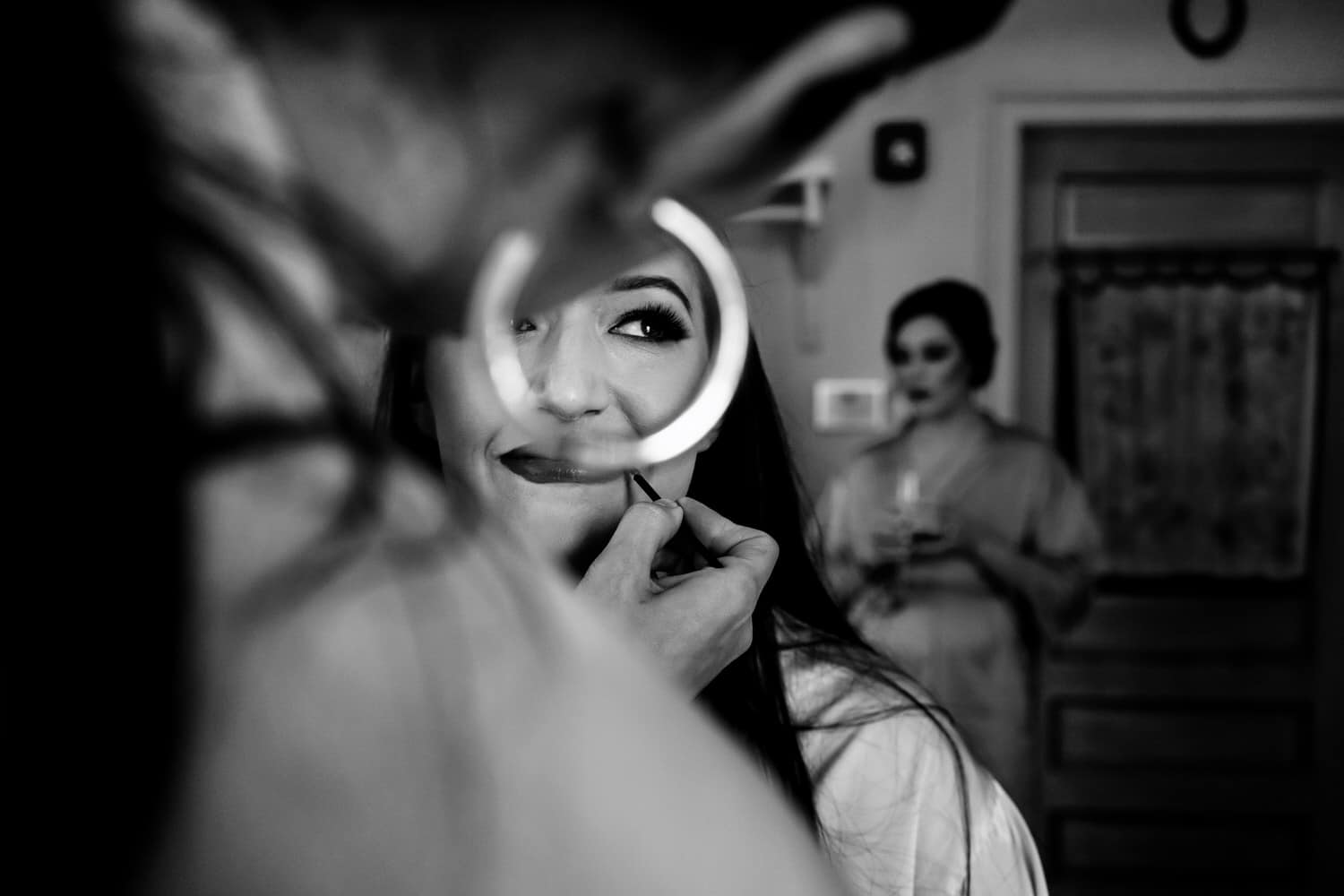 A candid black and white picture of a bride smiling, her eye visible through the hoop of a makeup artists earring. 