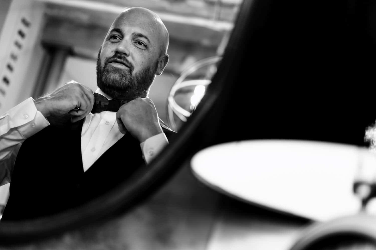 A candid black and white picture taken in the mirror of a bald man adjusting his bow tie on the morning of his Kansas City wedding at The Bride and The Bauer. 