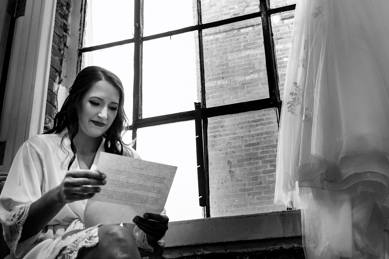 A candid black and white picture of a bride reading a letter from her groom, the edge of her wedding gown visible beside her on the morning of a summer wedding in Kansas City at The Bride and The Bauer. 