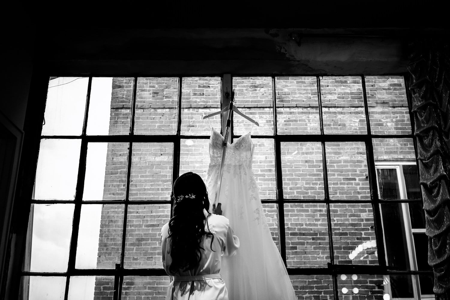 A candid black and white picture of a bride using a pole to pull her wedding gown down from a high window. 