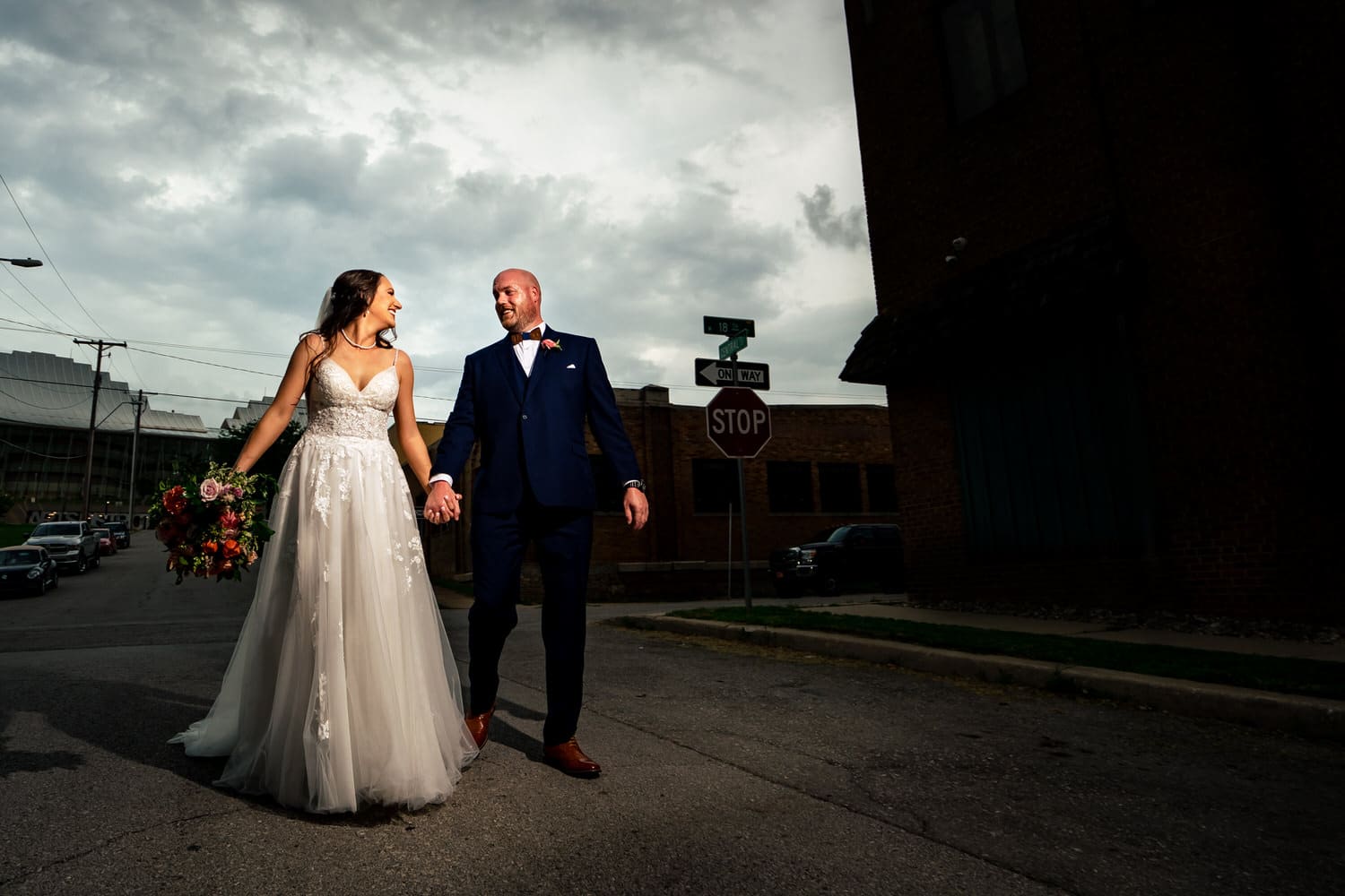 A candid, colorful picture of a bride and groom walking hand-in-hand down a city street, a stormy cloud visible behind them on their wedding day. 