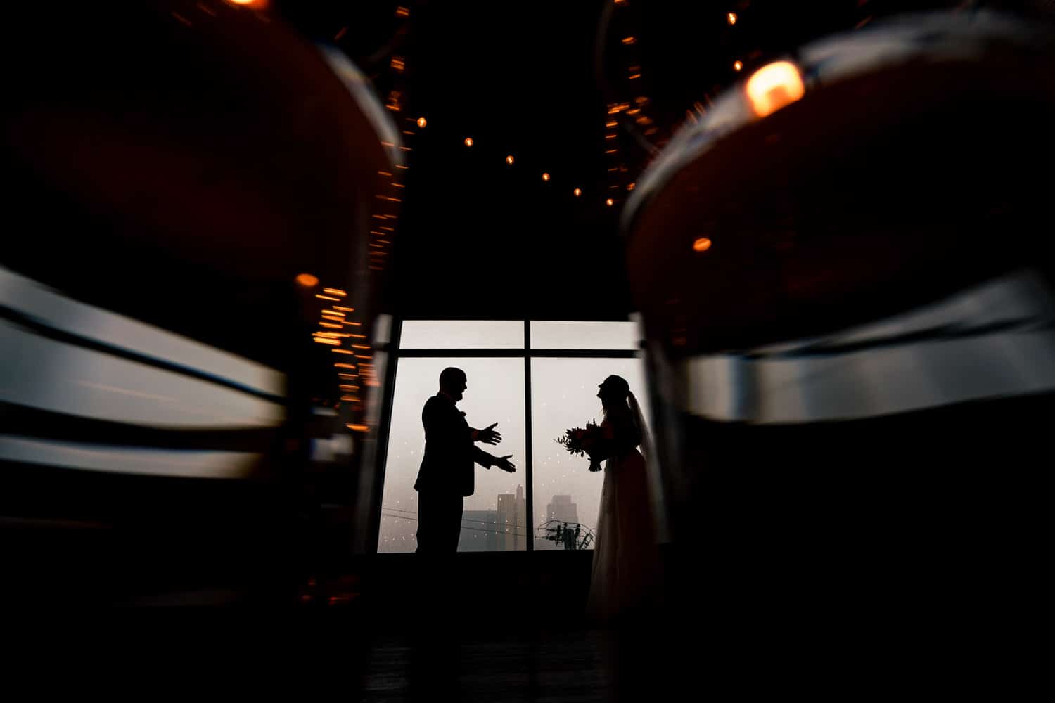 A silly, candid silhouette of a groom holding his arms out to his bride, taken between two lit candles, with the Kansas City skyline visible behind them. 