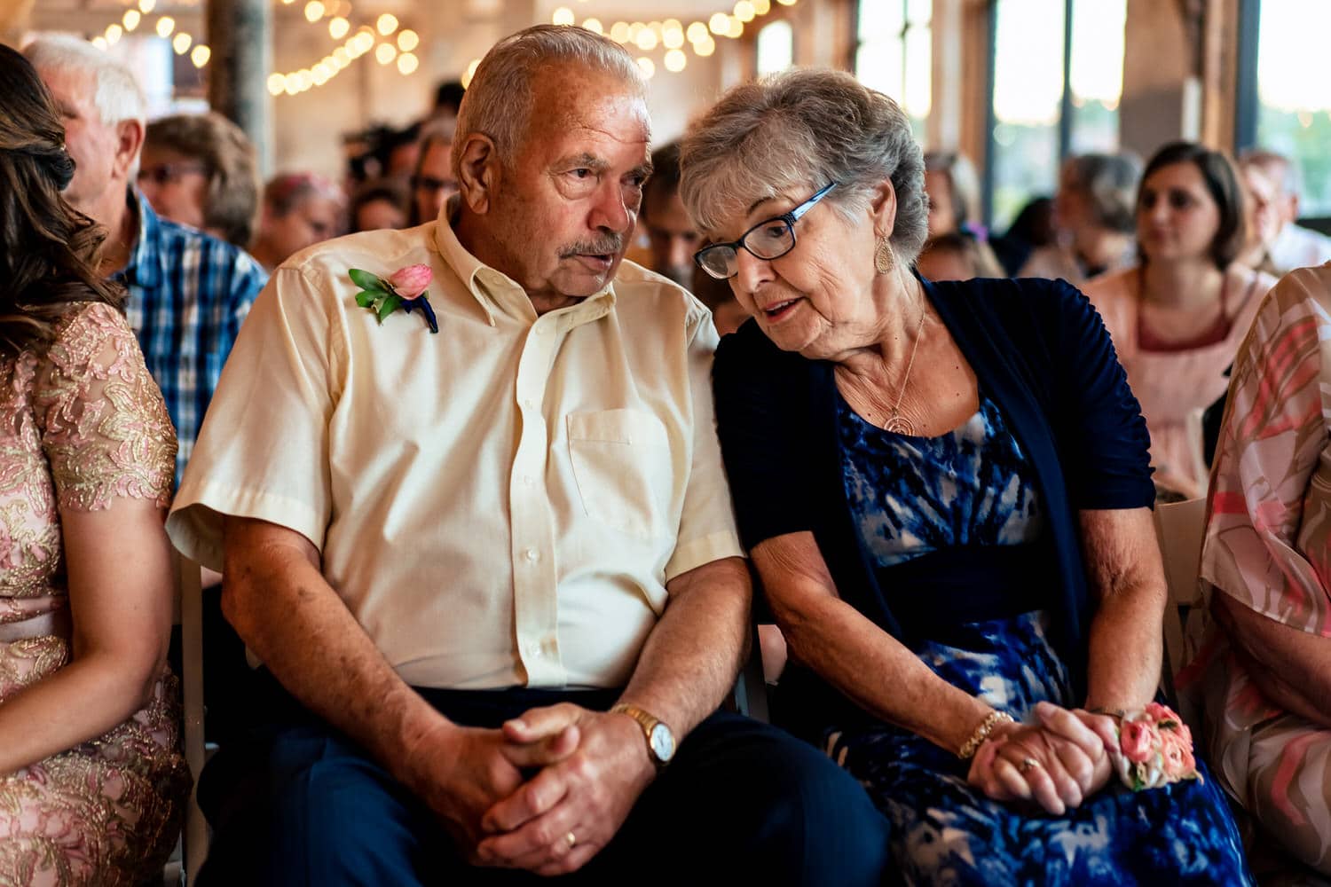A colorful, candid picture of a bride's grandparents whispering to each other as they wait for the bride to walk down the aisle during a summer wedding at The Bride and The Bauer in Kansas City. 