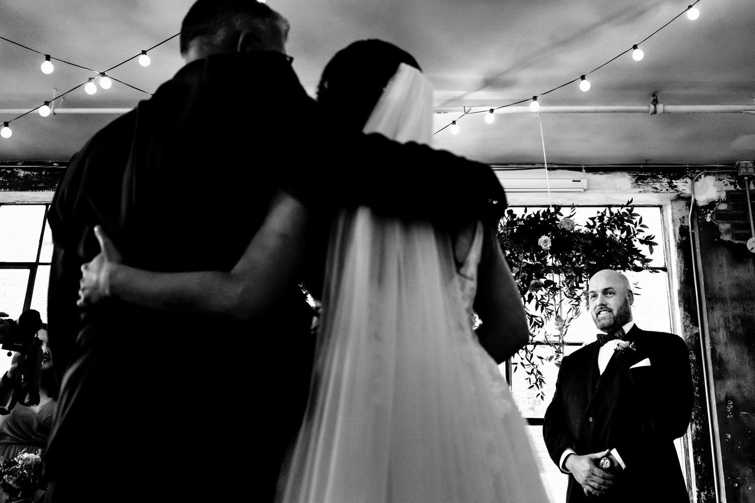 A candid black and white picture taken from behind the bride, of her dad hugging her as her groom smiles. 