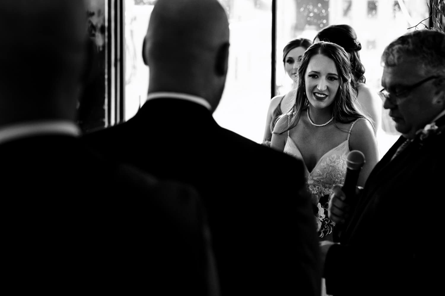 A candid black and white picture of a bride smiling at her groom as he reads his wedding vows to her during their summer wedding ceremony at The Bride and the Bauer in Kansas City. 