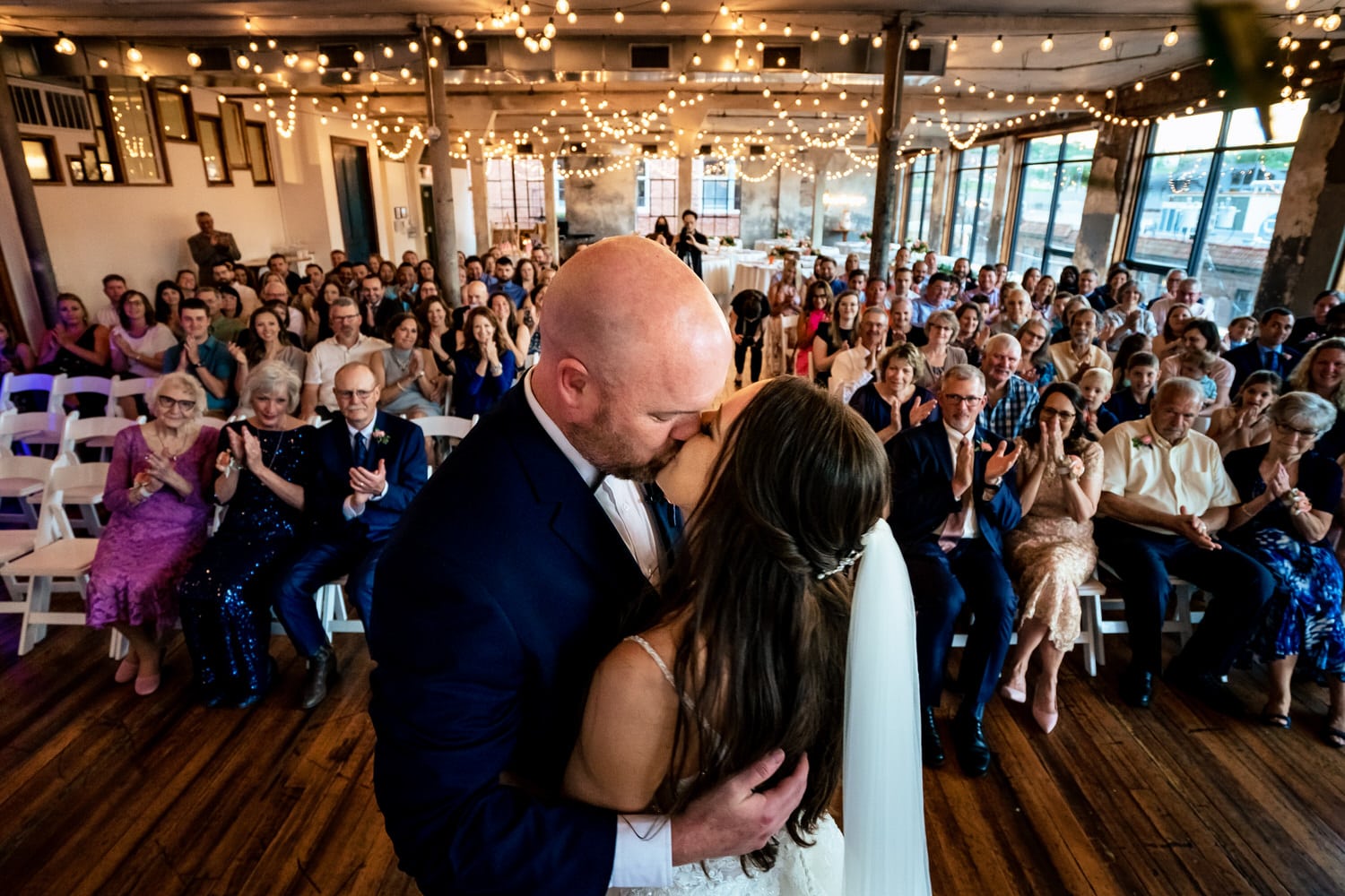 A colorful, candid picture taken from behind the bride and groom, looking back at the audience as they share their first kiss; their guests clapping and cheering in the background. 