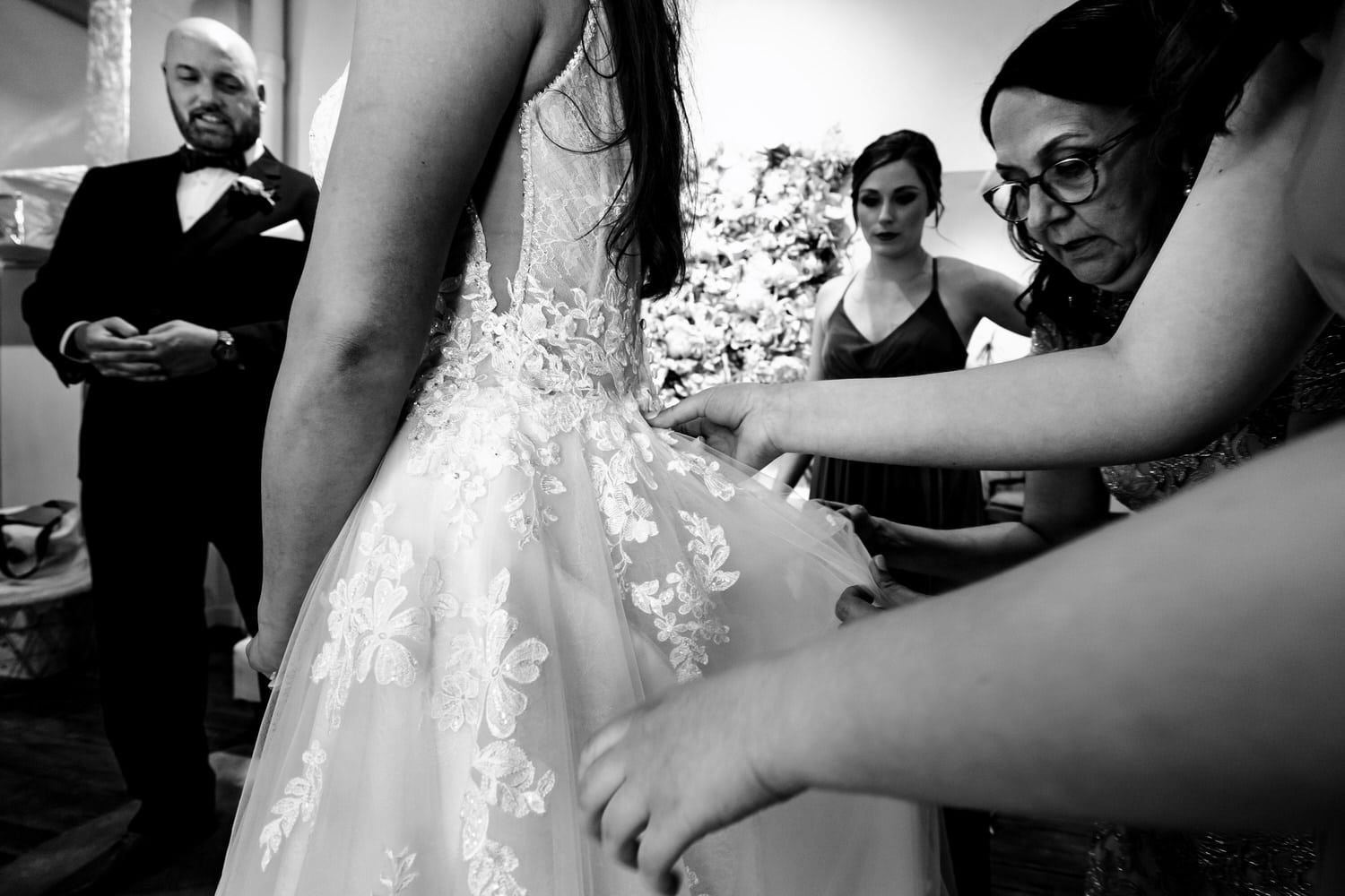 A candid black and white picture of a bride's mom and sister bustling her wedding gown. 