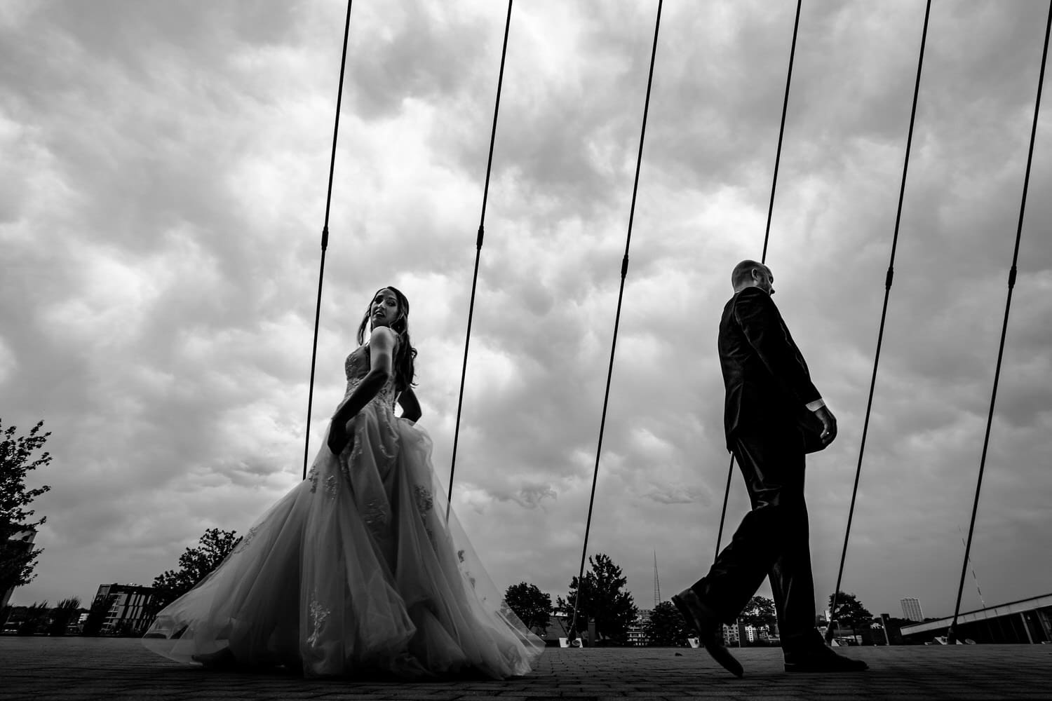 A candid black and white picture of a bride holding her gown, walking away from her groom and looking back over her shoulder; stormy skies visible behind them. 
