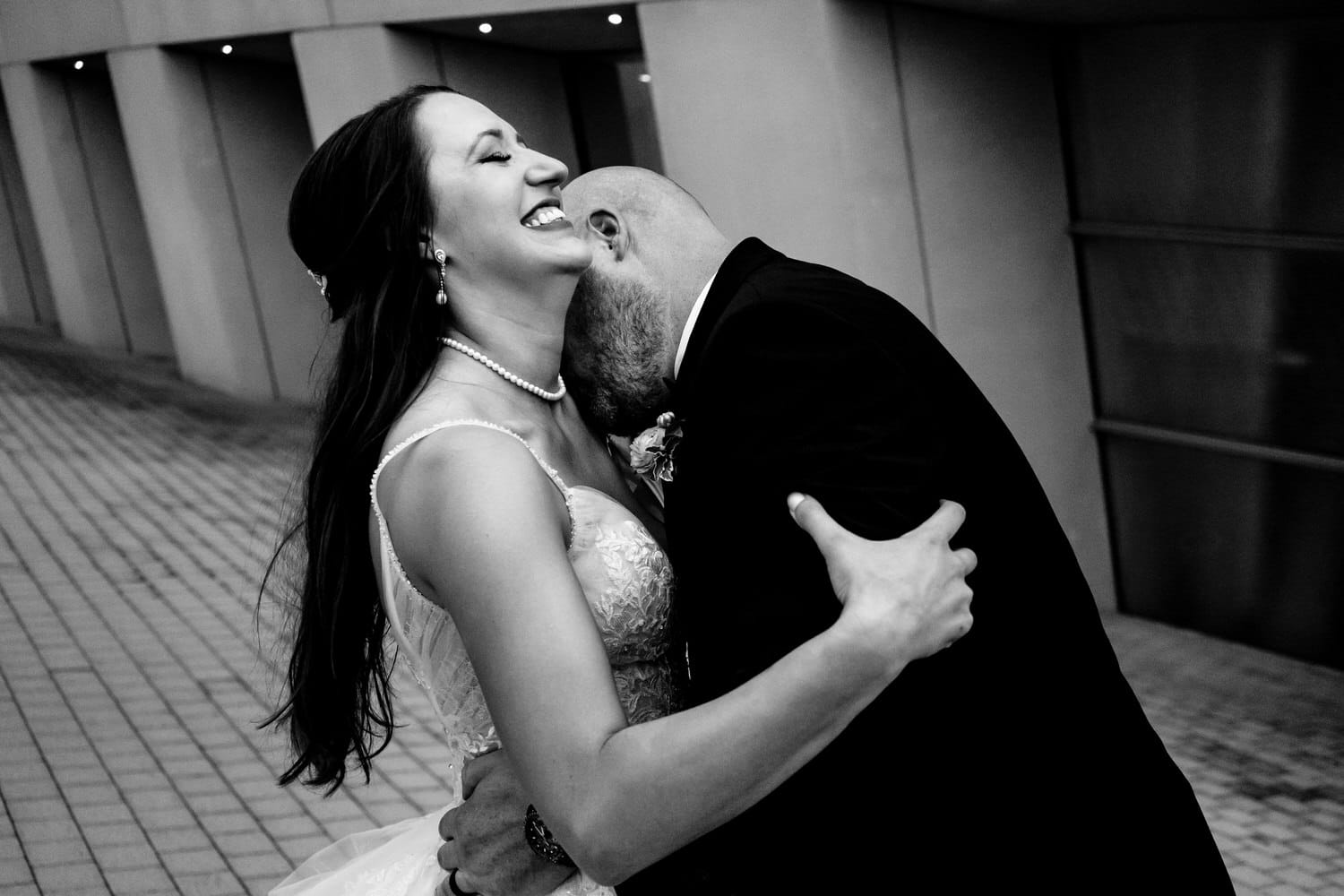 A candid black and white picture of a groom kissing his bride's neck as she laughs hysterically on their Kansas City wedding day. 