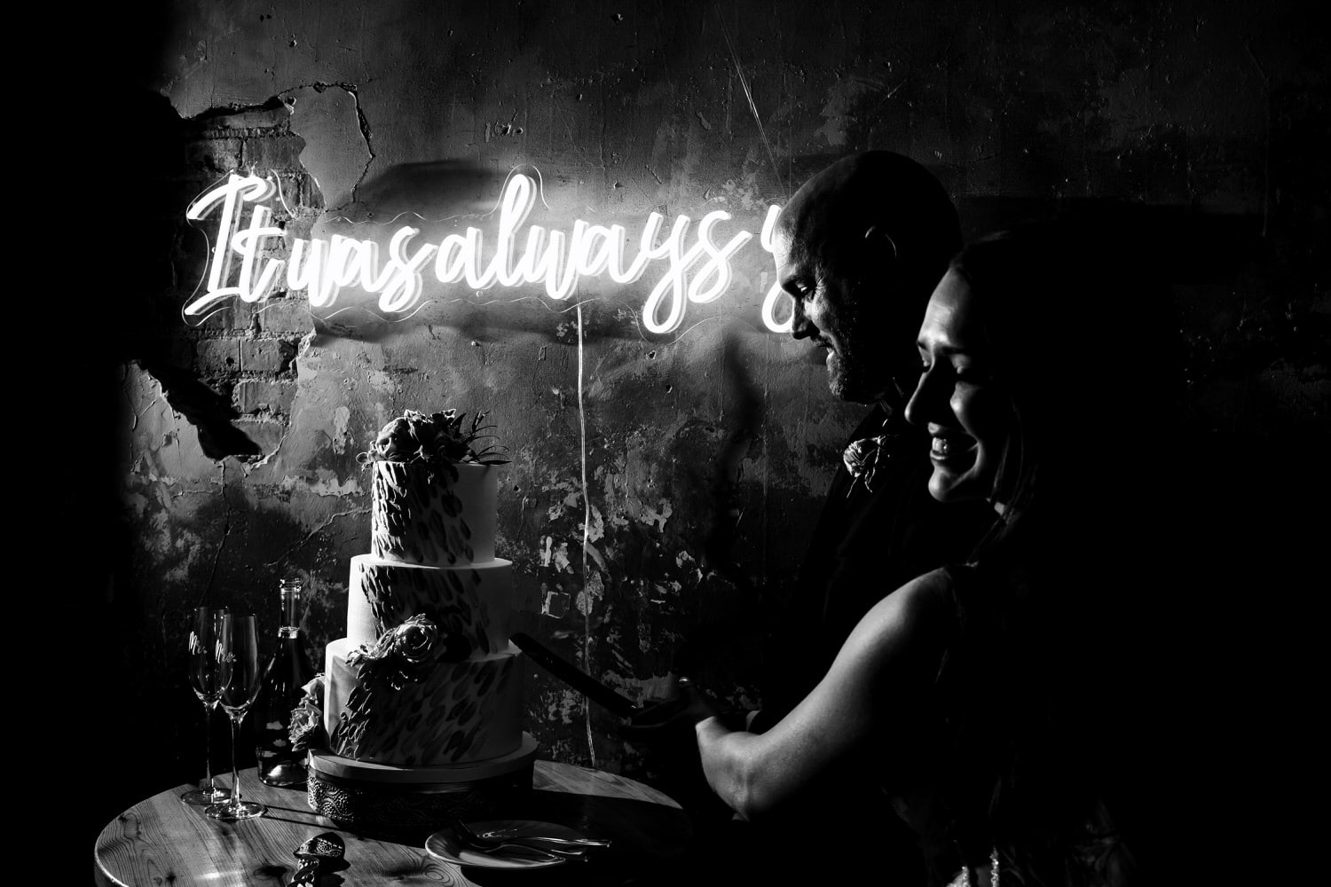 A candid black and white picture of a bride and groom leaning in to cut their wedding cake, an "it was always you" neon sign visible behind them. 