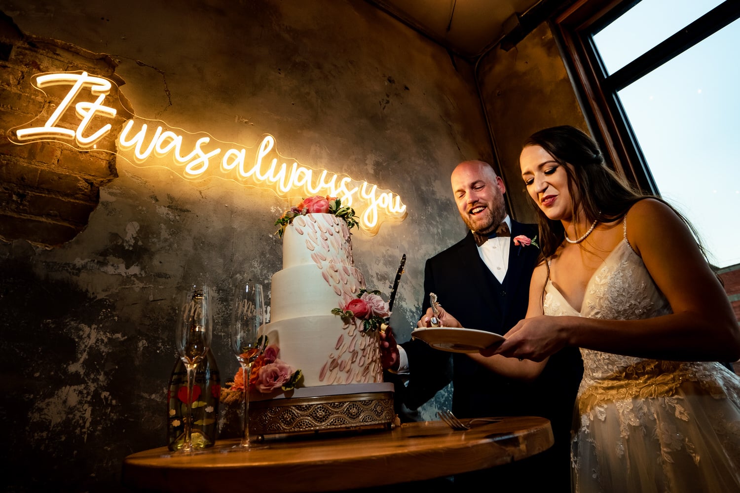A colorful, candid picture of a bride and groom getting ready to feed each other wedding cake, a neon, "it was always you" sign visible behind them. 