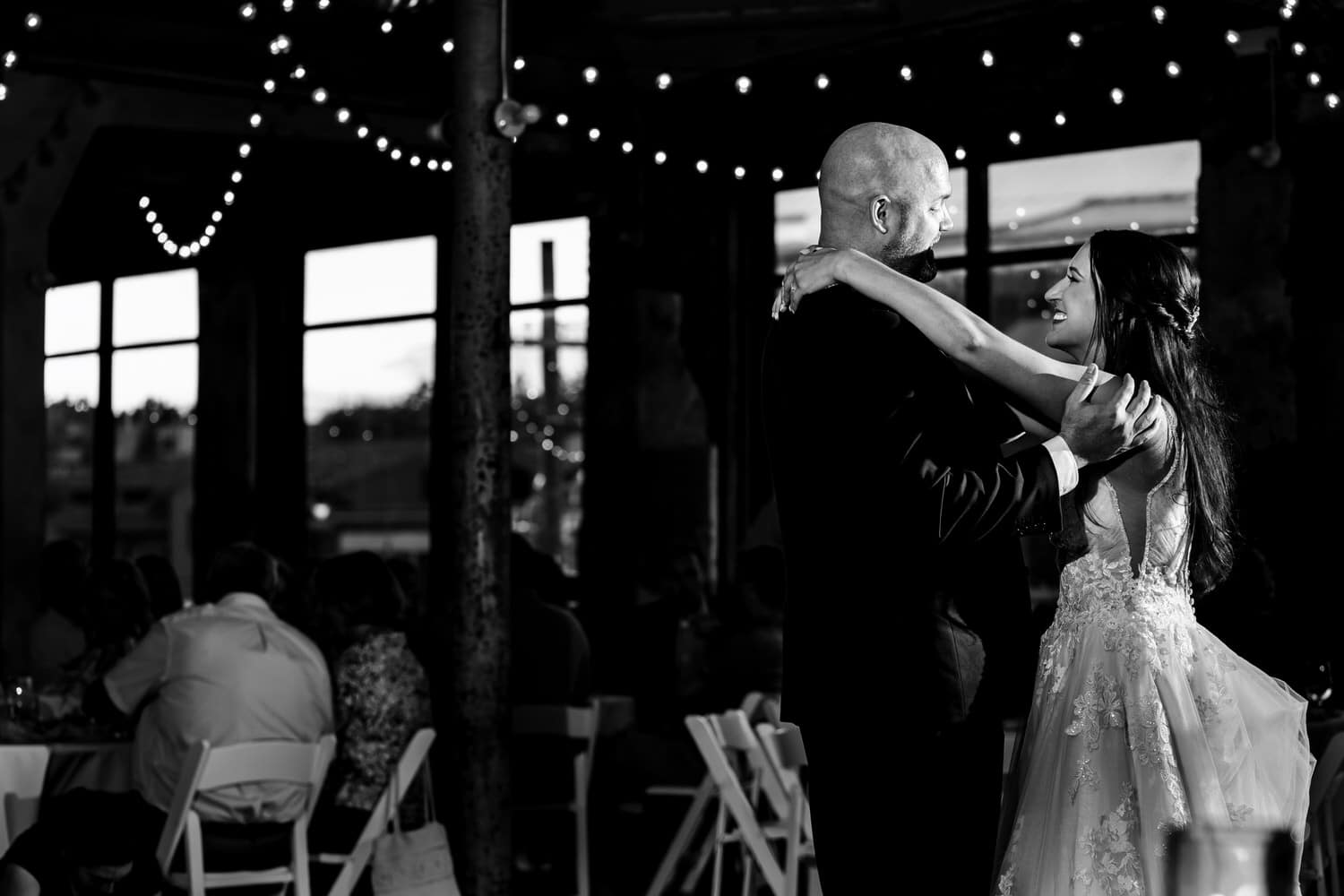 A wide picture of a bride and groom dancing together on a dance floor, lines of string lights visible above them during their wedding reception at The Bride and The Bauer in Kansas City. 