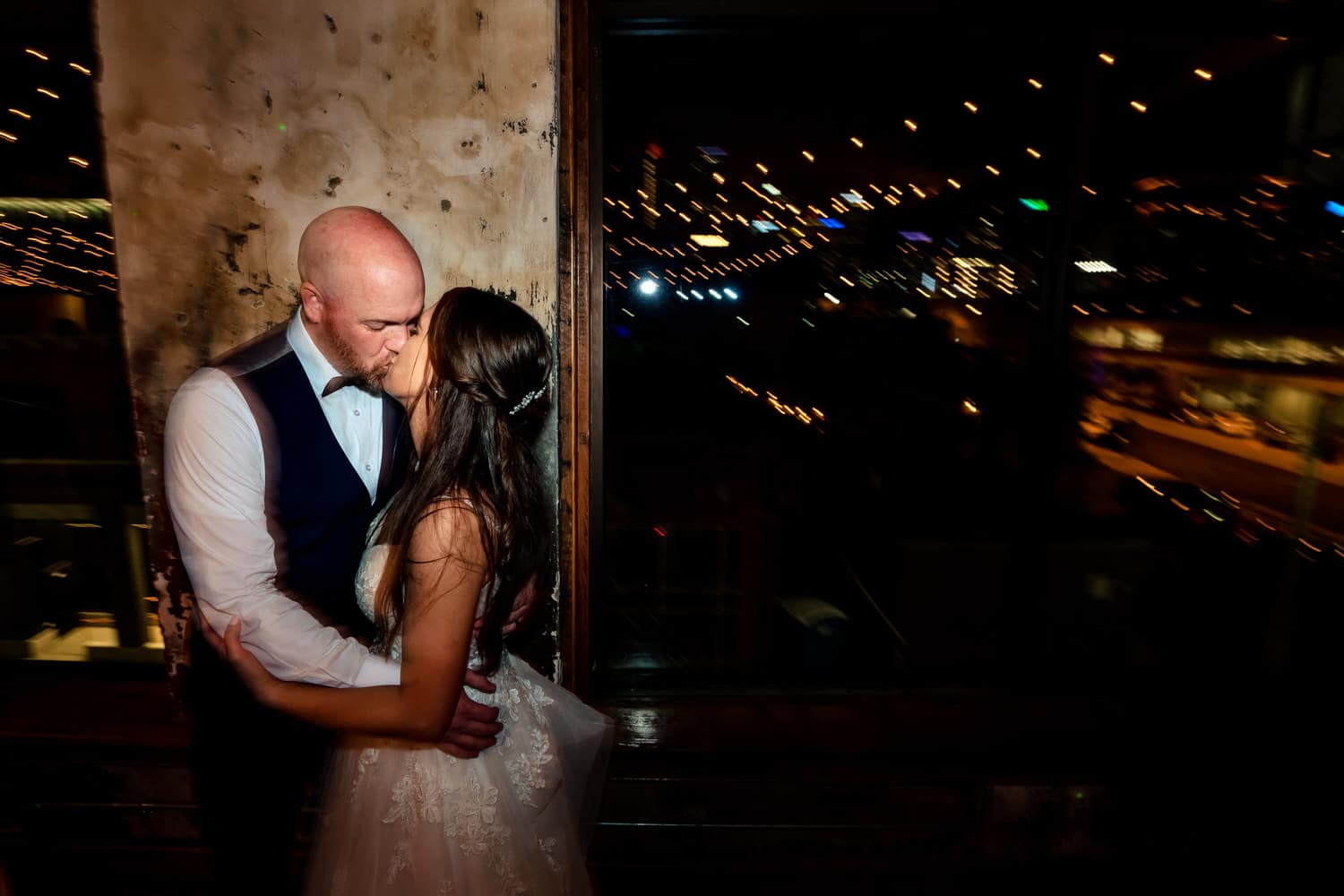 A candid picture of a bride and groom sharing a kiss, the twinkling city lights visible behind them during their wedding reception at The Bride and The Bauer. 
