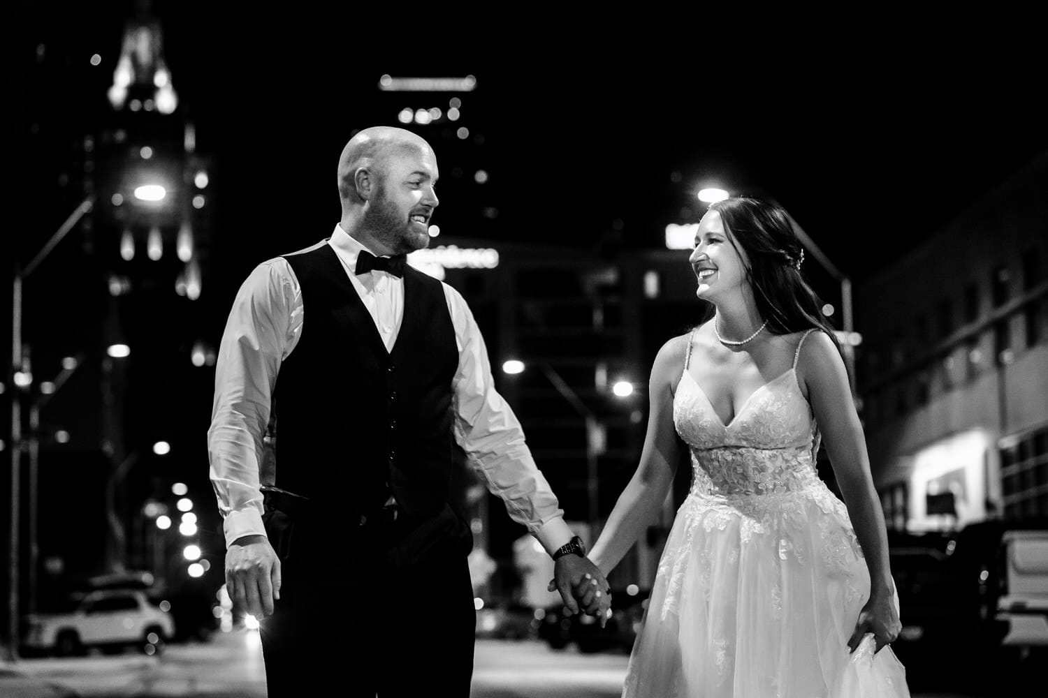 A candid black and white picture of a bride and groom holding hands, walking down a city street, the Kansas City skyline visible behind them on their wedding day. 