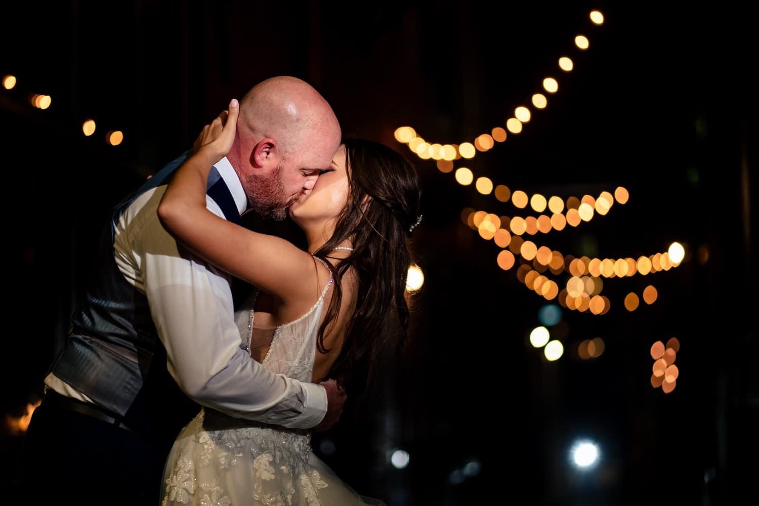 A wide, colorful picture of a bride and groom sharing a kiss in an alley outside The Bride and The Bauer; a canopy of string lights visible above them. 