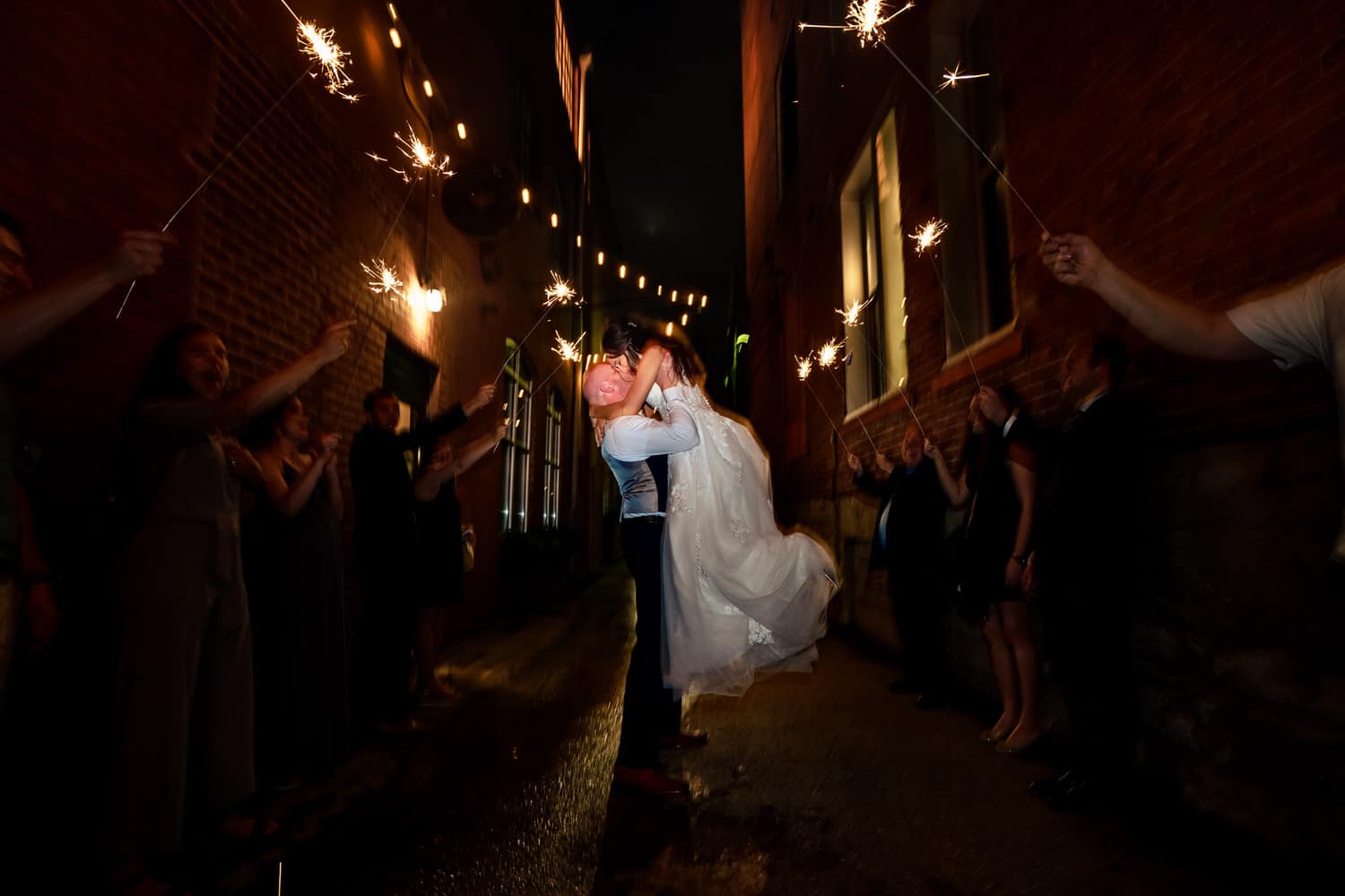 A candid picture of a groom lifting his bride into the air as family and friends wave sparklers around them during their wedding reception sparkler exit. 