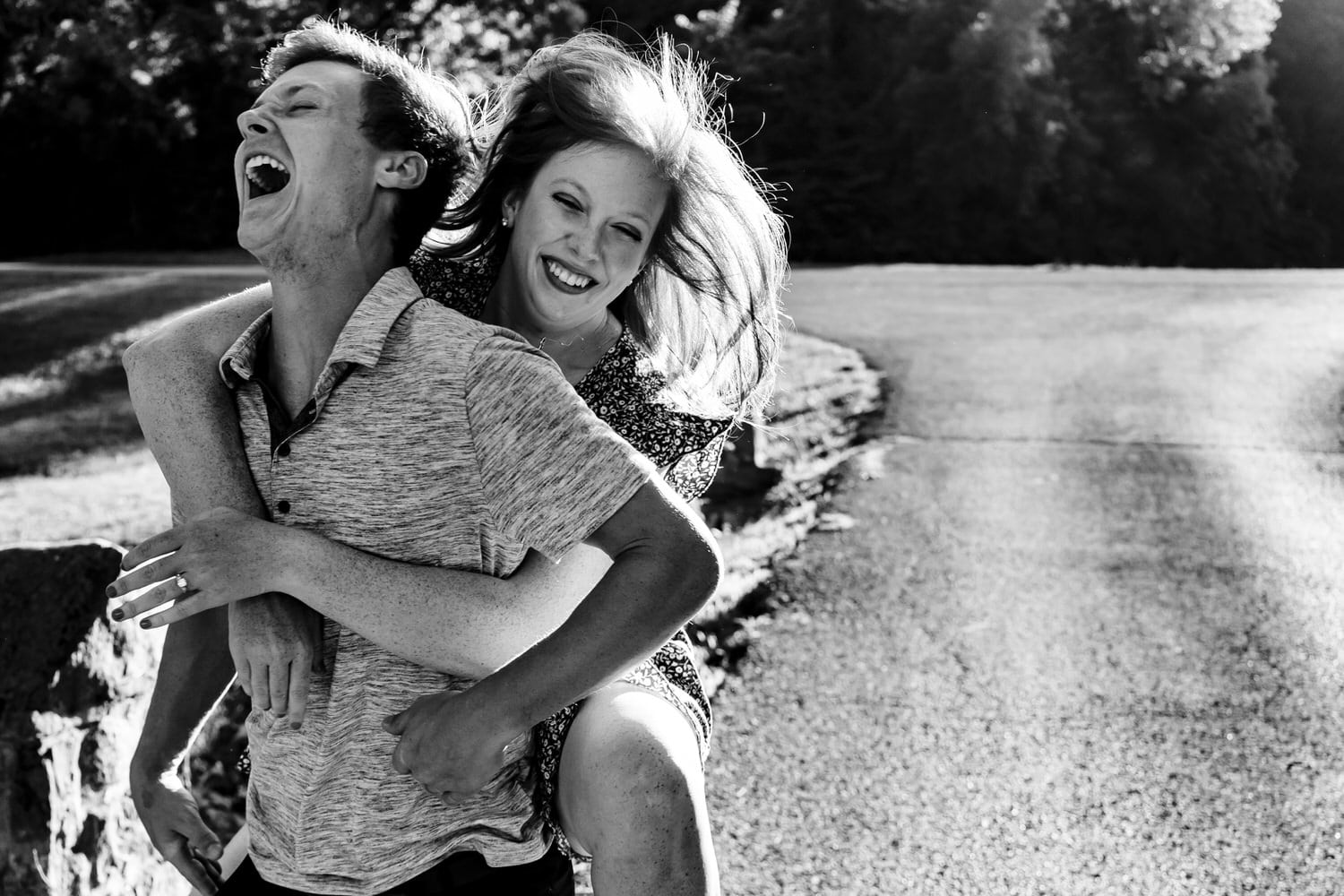 A candid black and white picture of an engaged couple laughing hysterically as he gives her a piggy-back ride during their summer engagement session at Wyandotte County Lake Park in Kansas City. 