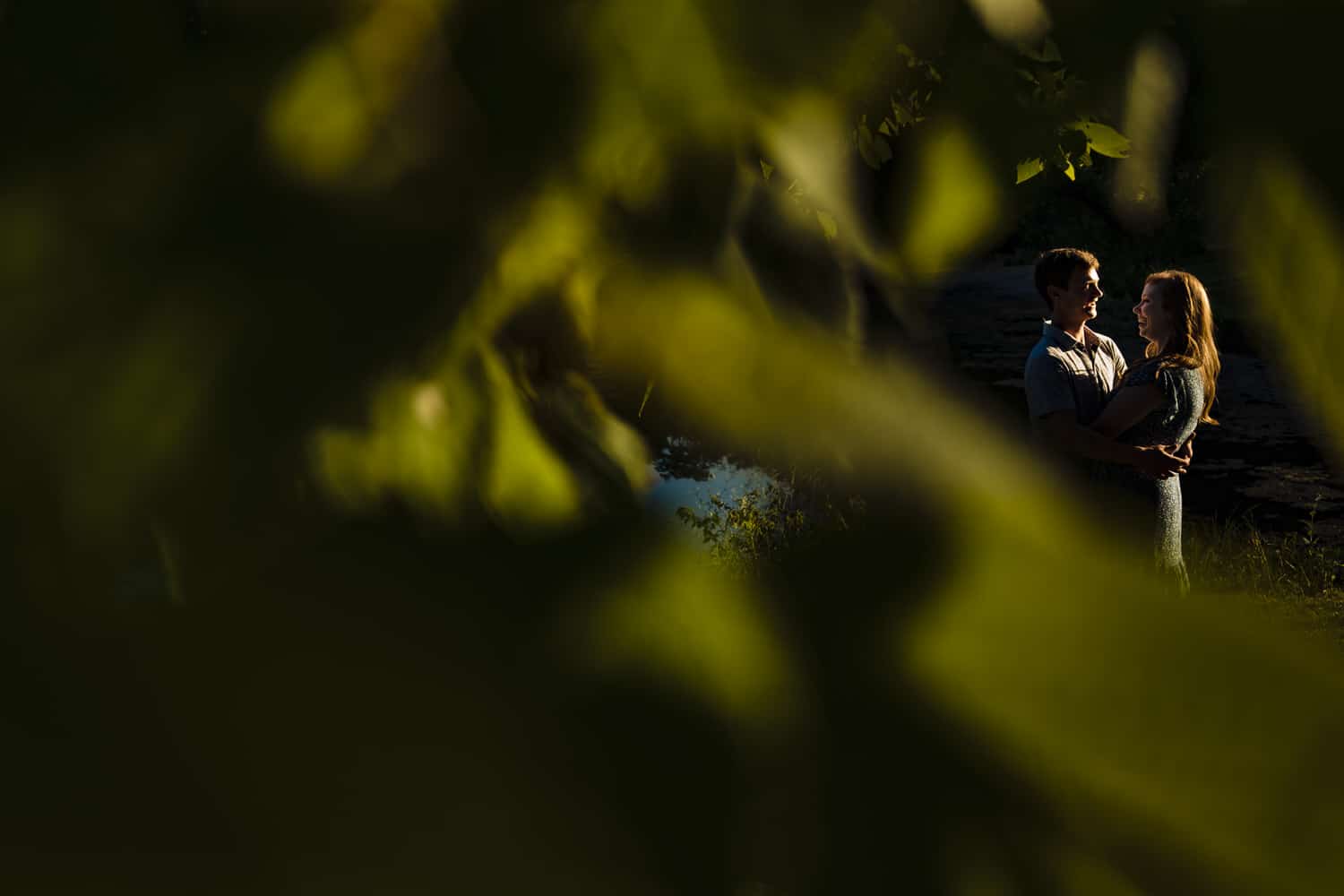 A candid picture taken through a branch of trees of an engaged couple sharing an embrace and laughing together during their engagement session in Kansas City. 