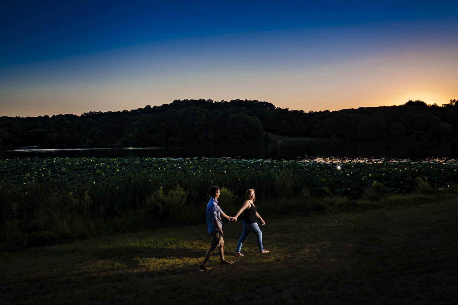 A candid picture of an engaged couple holding hands and walking across a lawn at sunset during their engagement session at Wyandotte County Park in Kansas City. 