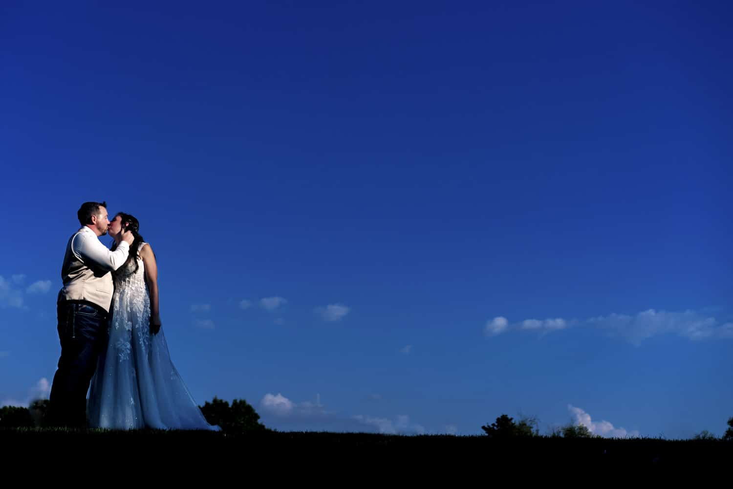 A colorful picture of a bride and groom standing against a background of bright blue sky, sharing a kiss on their summer wedding day at the Countryside Chalet in Kansas City. 