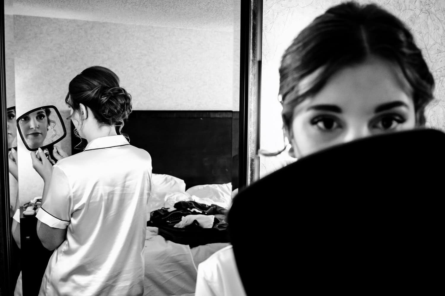 A candid black and white picture of a woman looking into a handheld mirror at the back of her hair in another mirror on the morning of her summer wedding in Kansas City. 