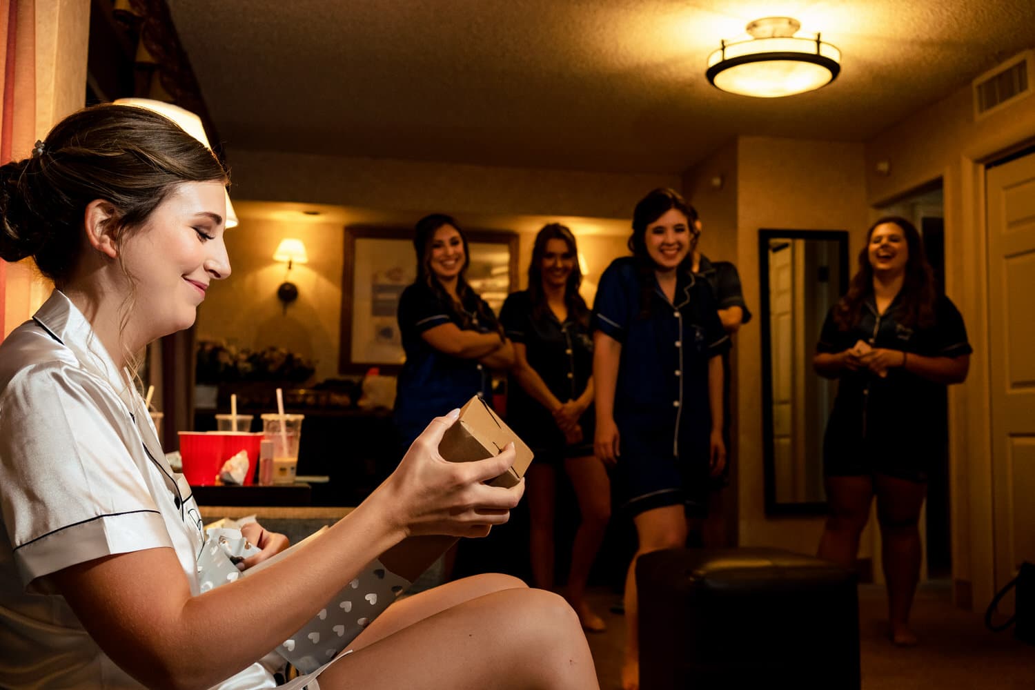 A candid picture of a bride in white pajamas opening a gift from her groom, her bridesmaids excitedly watching in the background. 