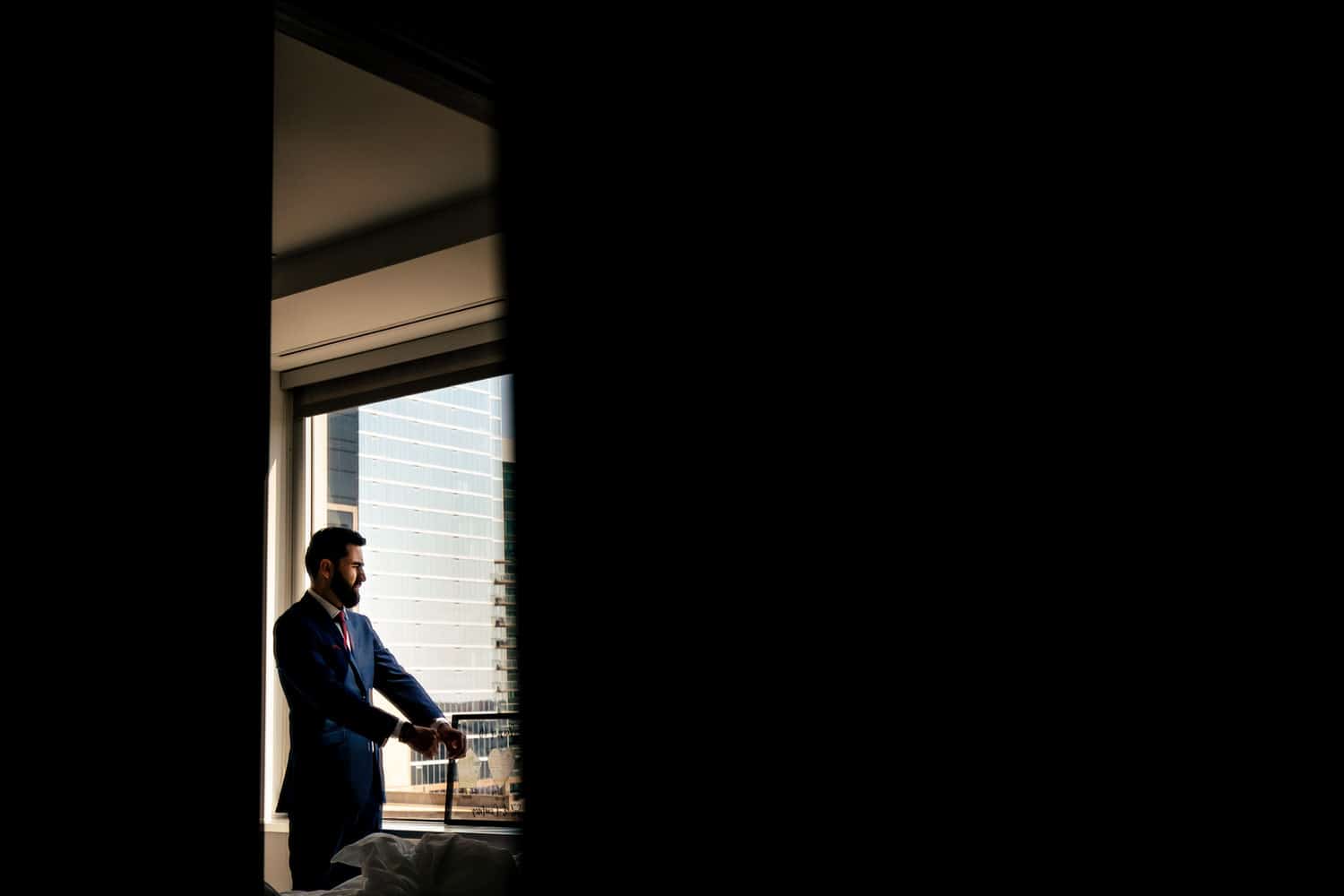 A candid picture taken through a doorway of a man adjusting his cuff links as he looks out a window at the downtown Kansas City skyline. 