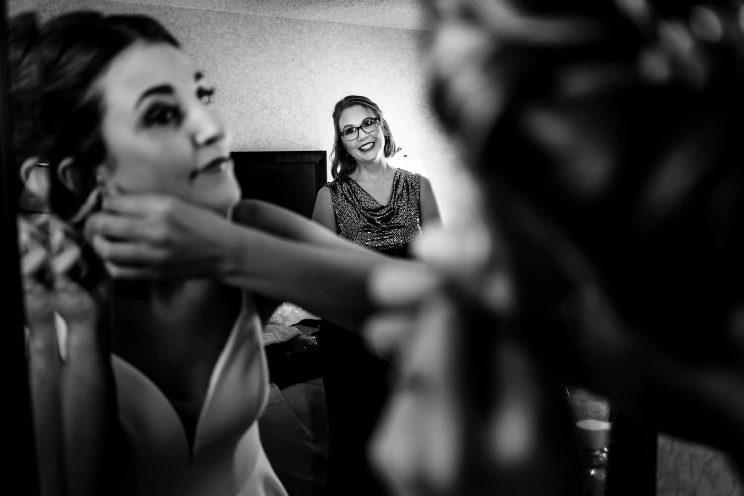 A candid black and white picture of a bride putting her earrings in, her mother visible in the background smiling at her. 