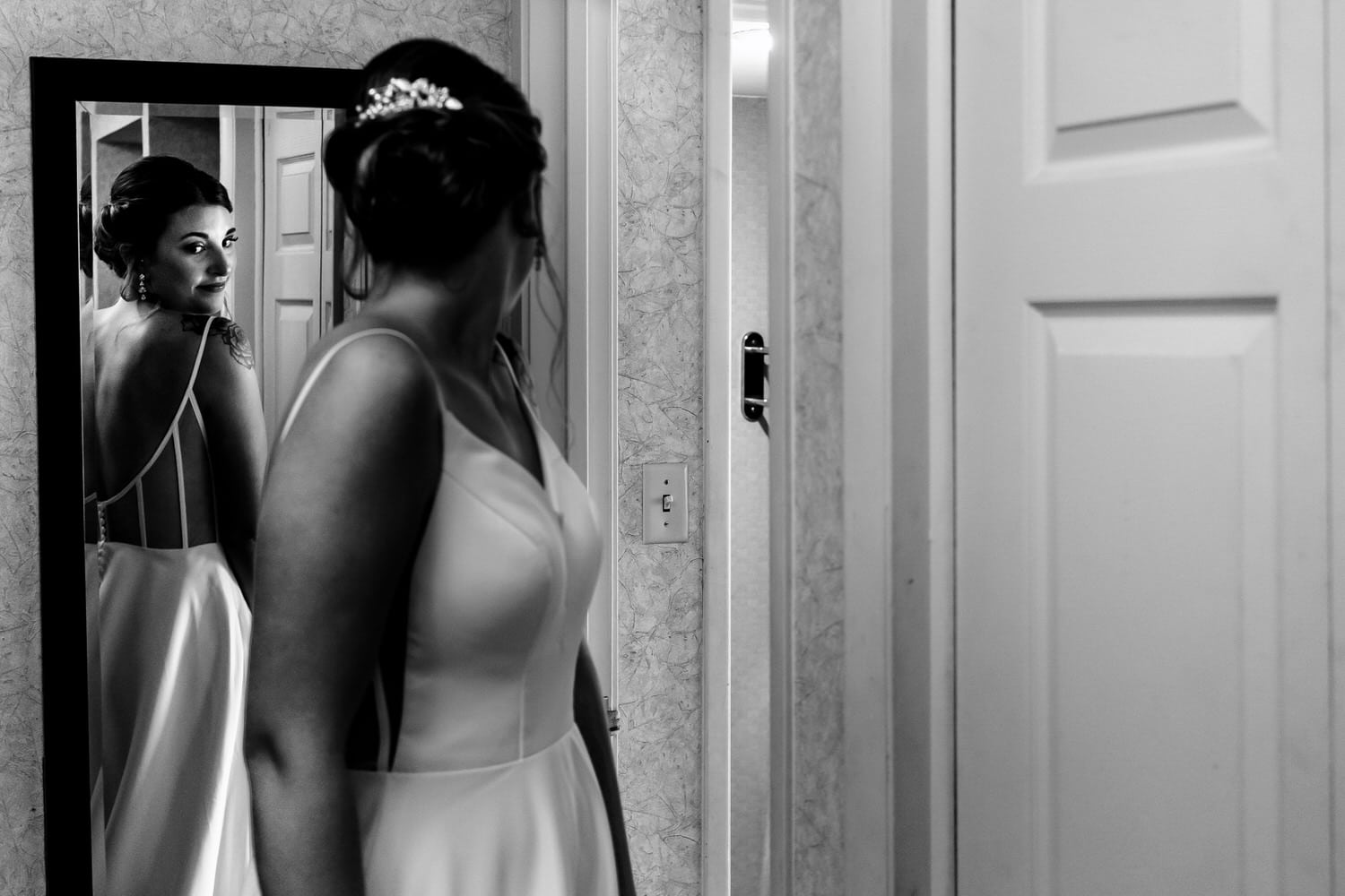 A candid black and white picture of a bride looking back into a mirror at the back of her wedding gown on a summer wedding day. 
